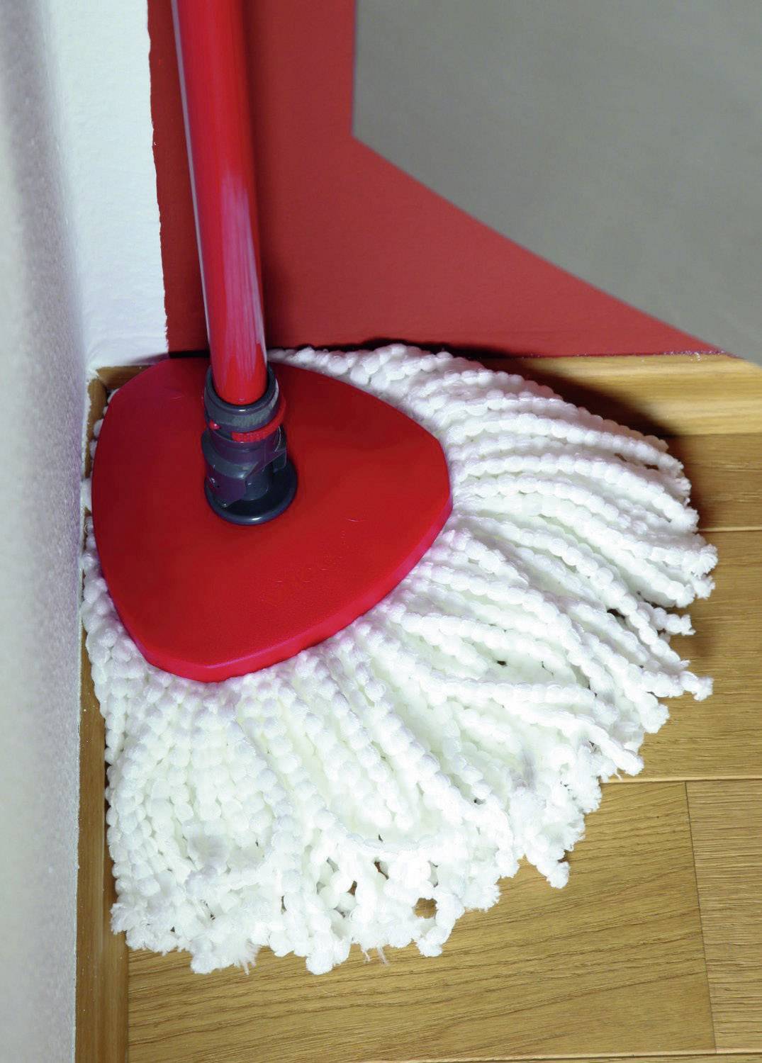 Červený mop leží v rohu na dřevěné podlaze.