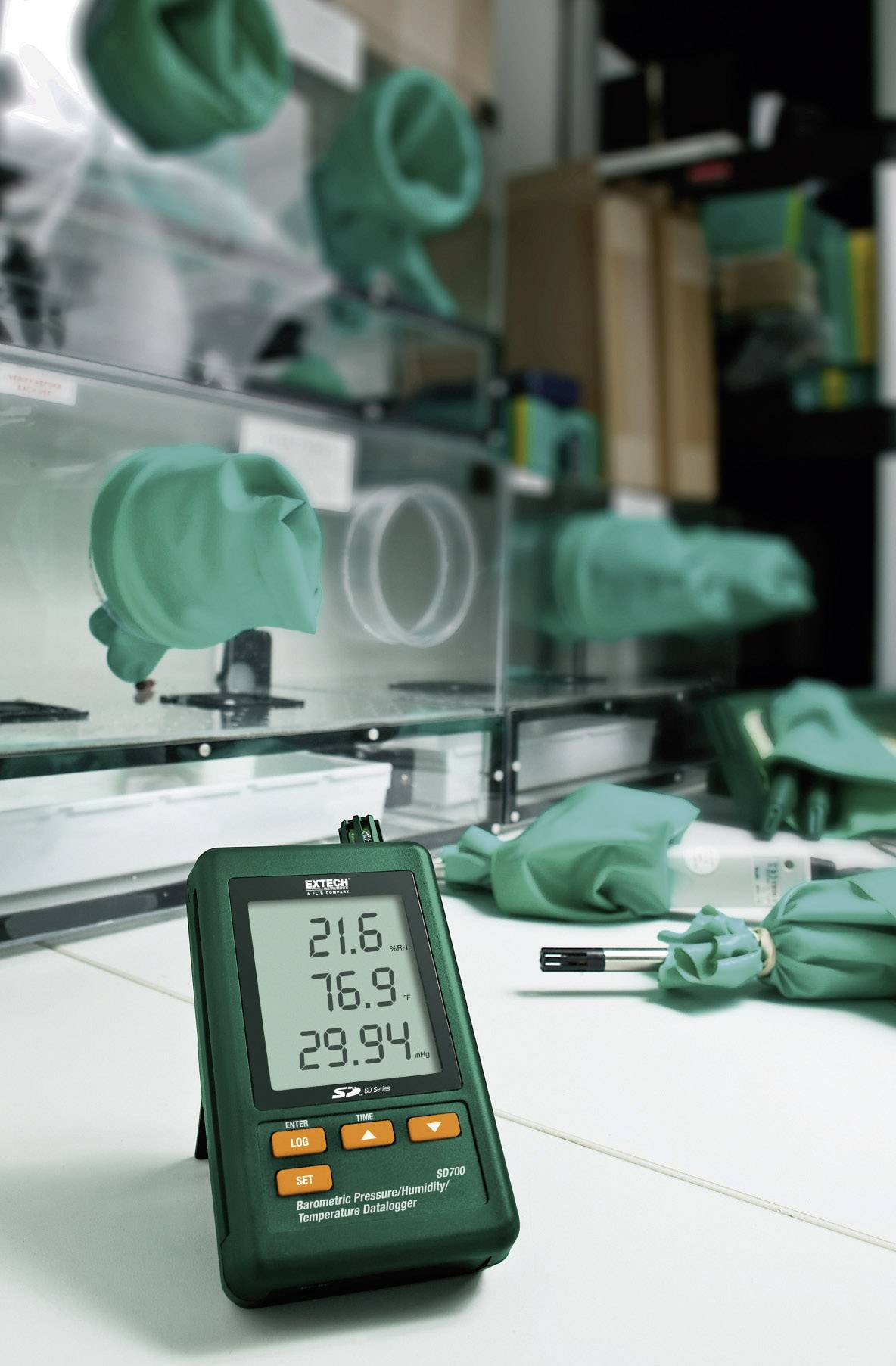 Digitální termo-hygrometr ukazuje teplotu na stole. V pozadí jsou laboratorní přístroje a zelené ochranné rukavice.