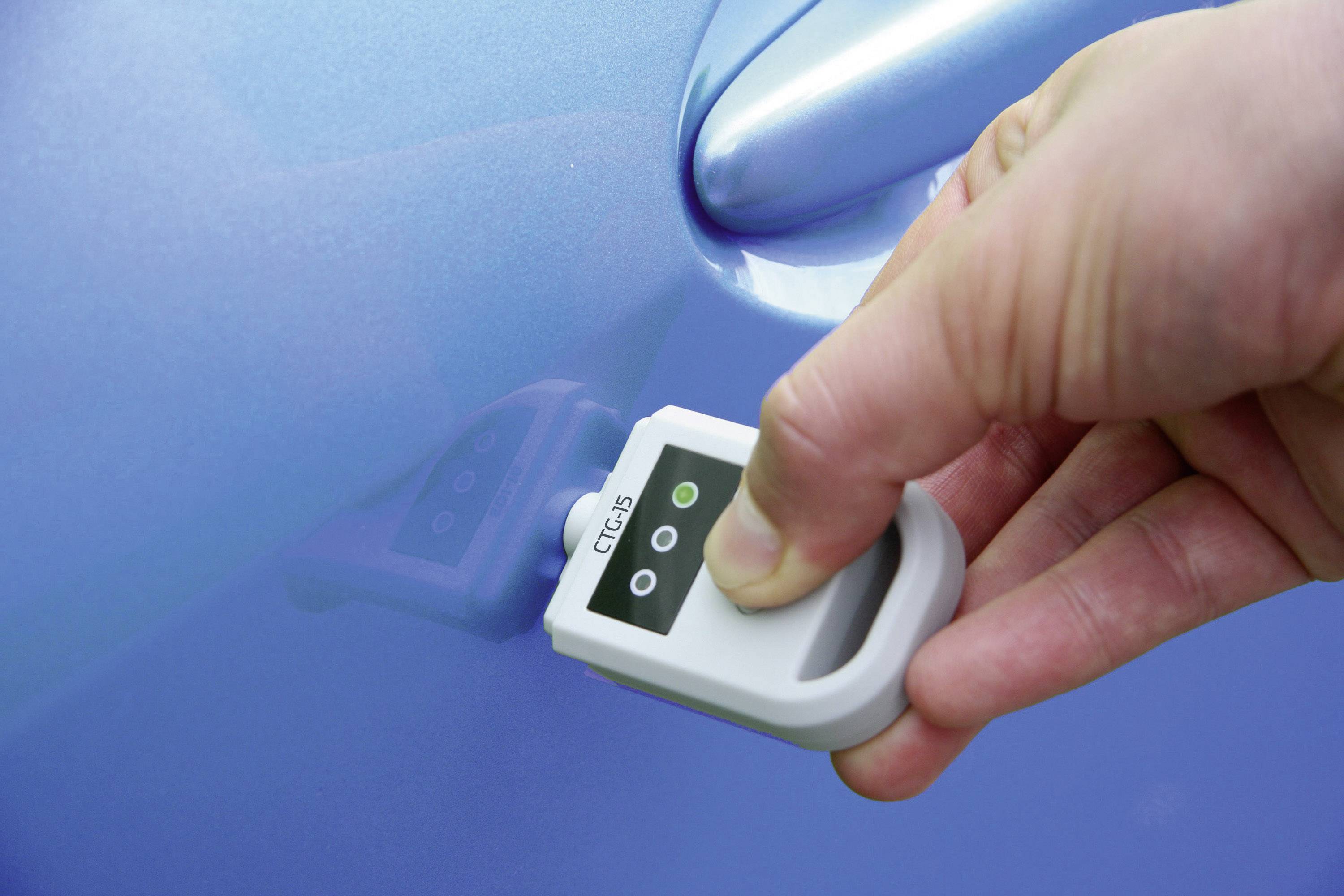 Jedna ruka drží měřicí přístroj přiložený k modré barvě automobilu, aby zkontrolovala tloušťku lakové vrstvy. Přístroj ukazuje zelený světelný indikátor.