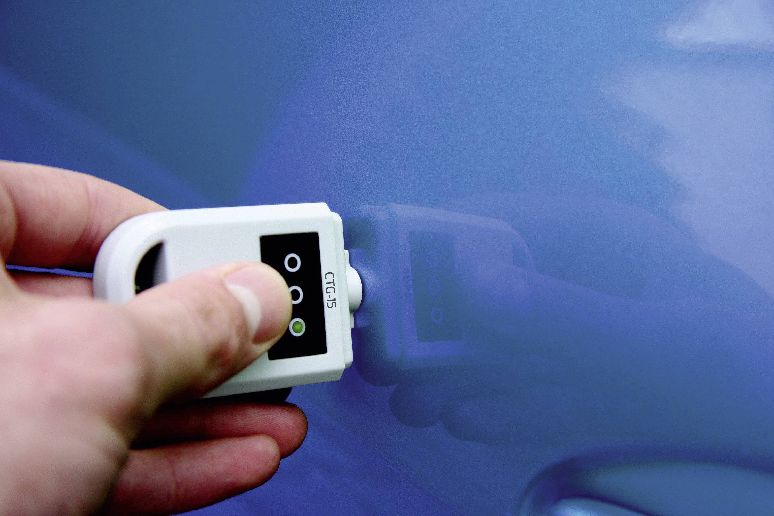 Jedna ruka drží přístroj na měření tloušťky laku na povrchu modrého automobilu, aby zkontrolovala tloušťku lakované vrstvy.