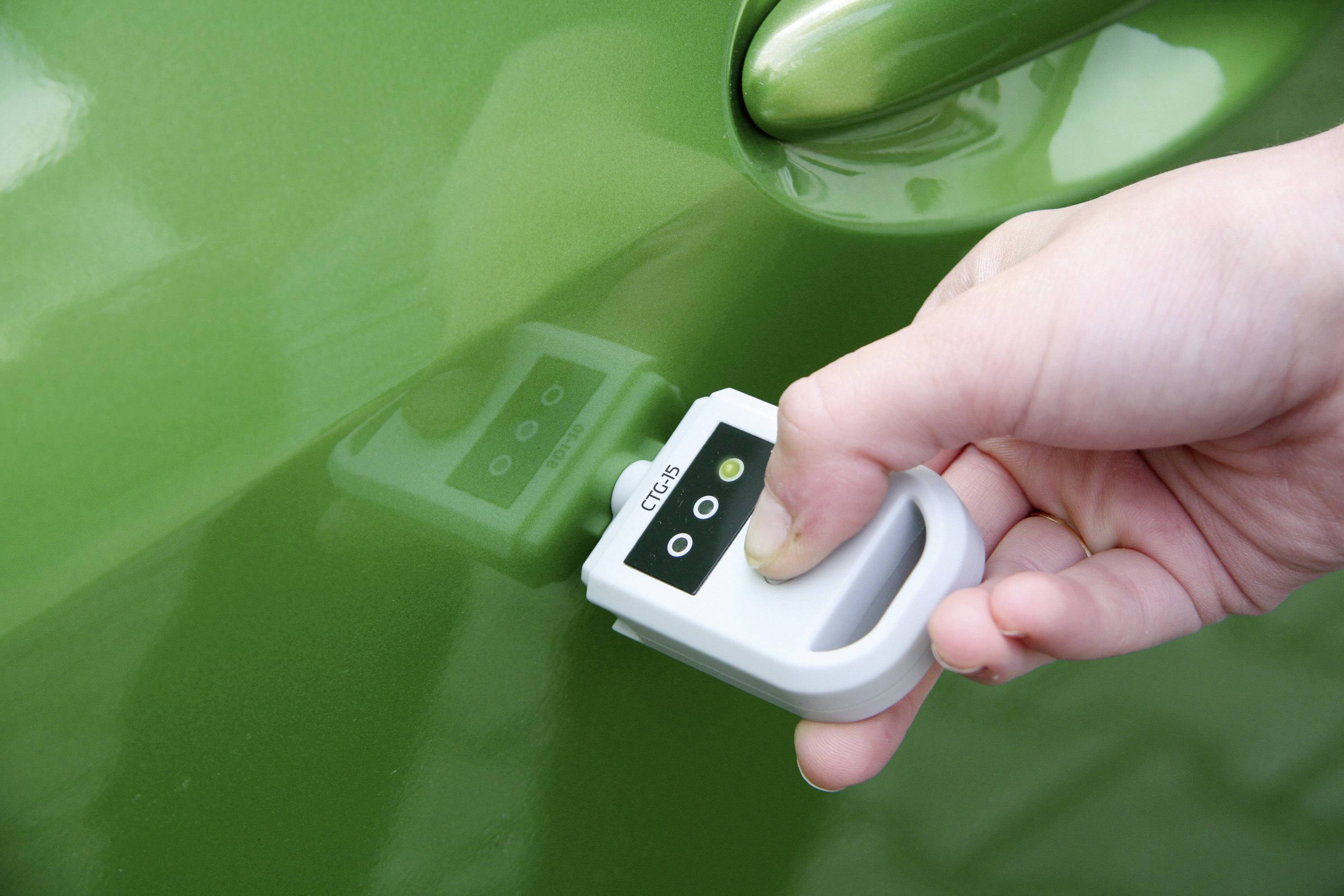 Ruka drží barevný měřicí přístroj u zeleného automobilu, aby zkontrolovala lak. Přístroj zobrazuje digitální hodnotu na malém displeji.