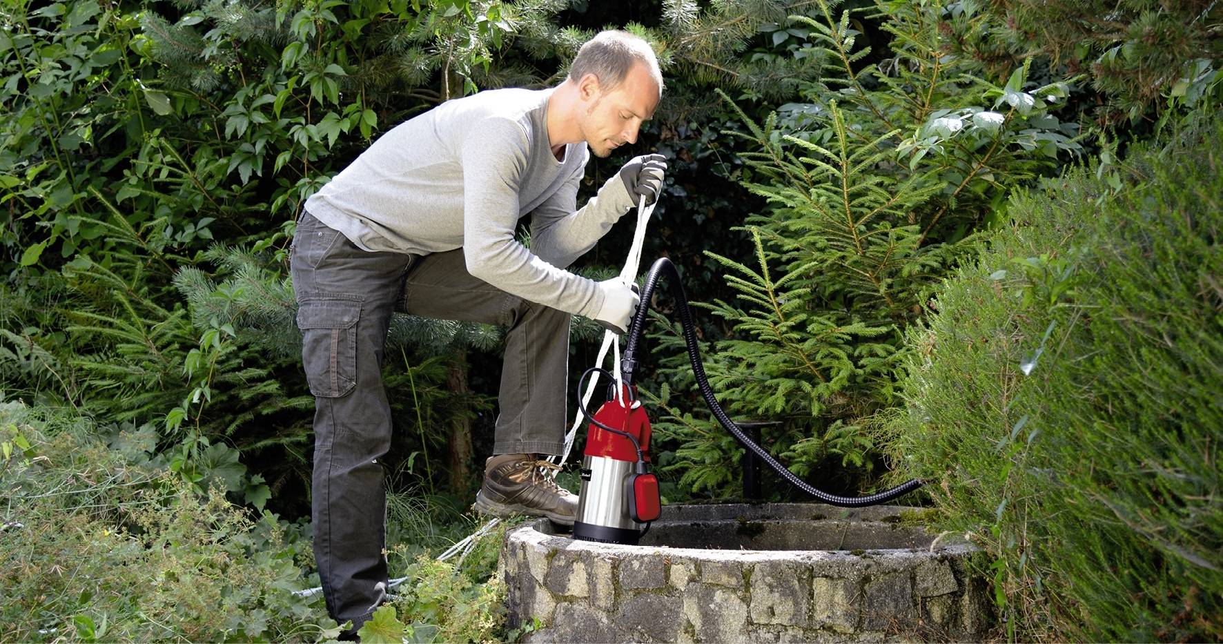 Osoba čerpá vodu z studny pomocí červené elektrické pumpy, obklopena hustou vegetací a keři.