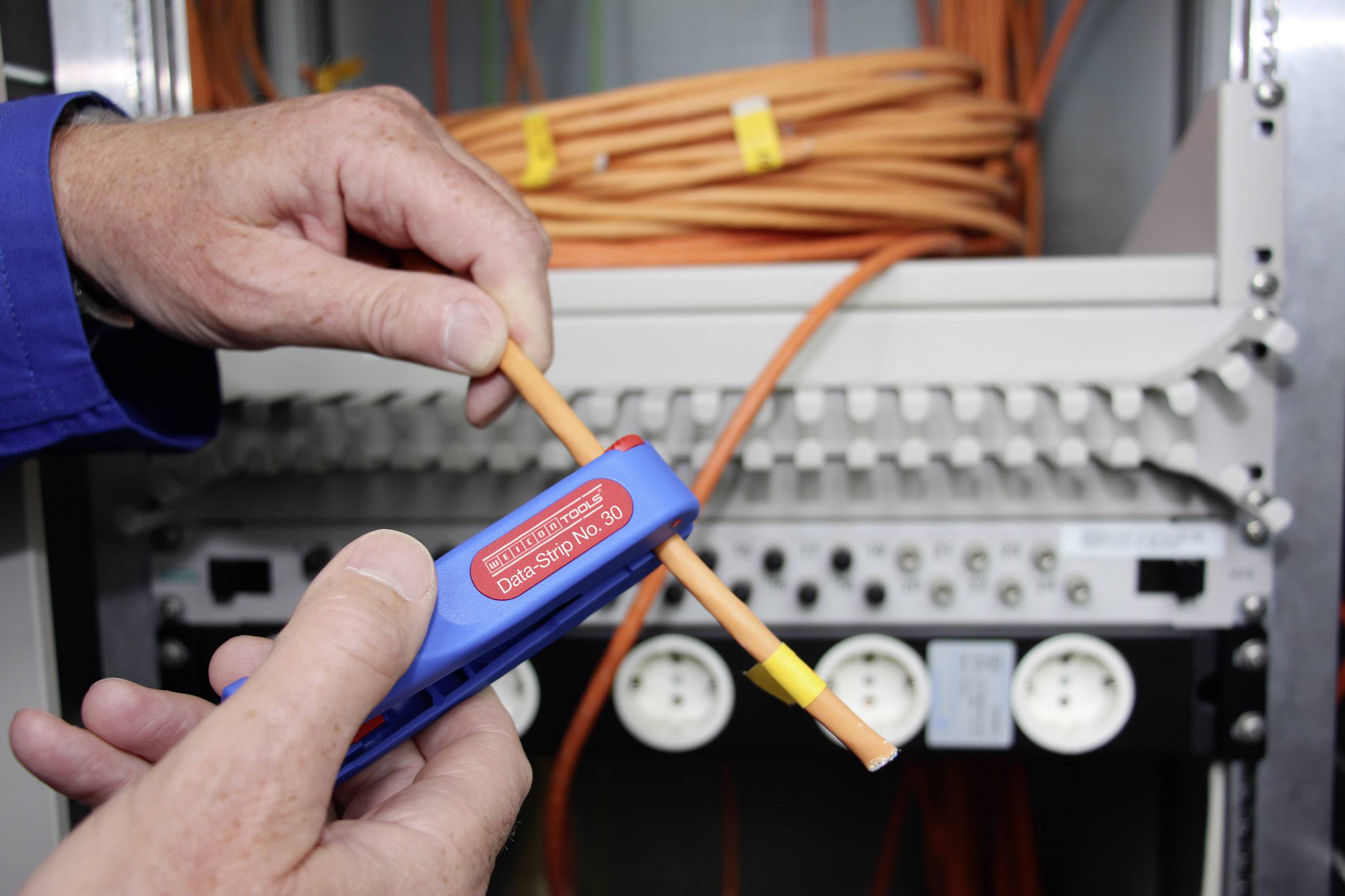 Osoba drží odizolovací nástroj, aby odhalila oranžový kabel. V pozadí jsou vidět síťové konektory.