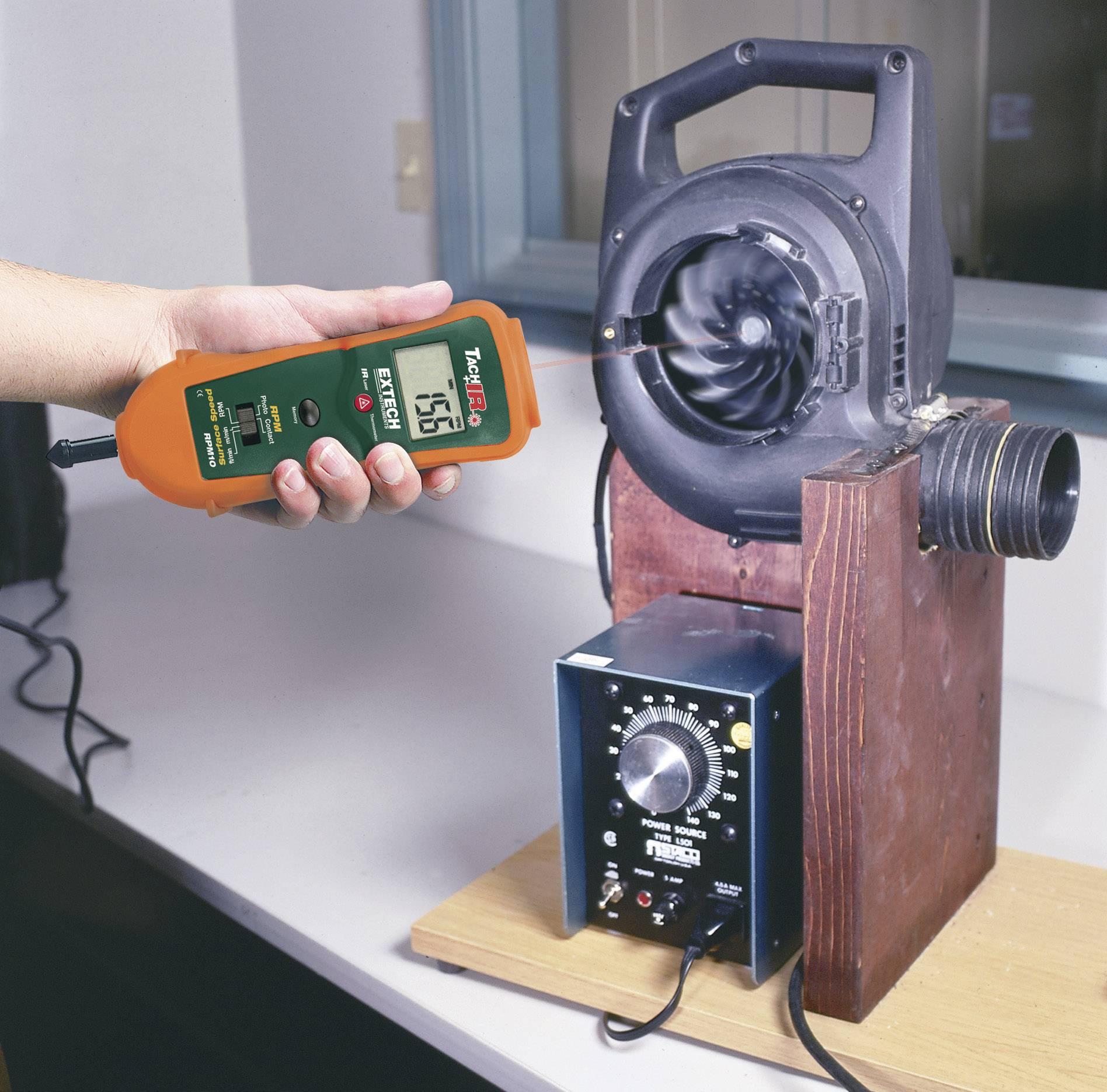 Jedna osoba měří proudění vzduchu ventilátoru pomocí anemometru v laboratoři. Anemometr ukazuje hodnotu '7,56'.