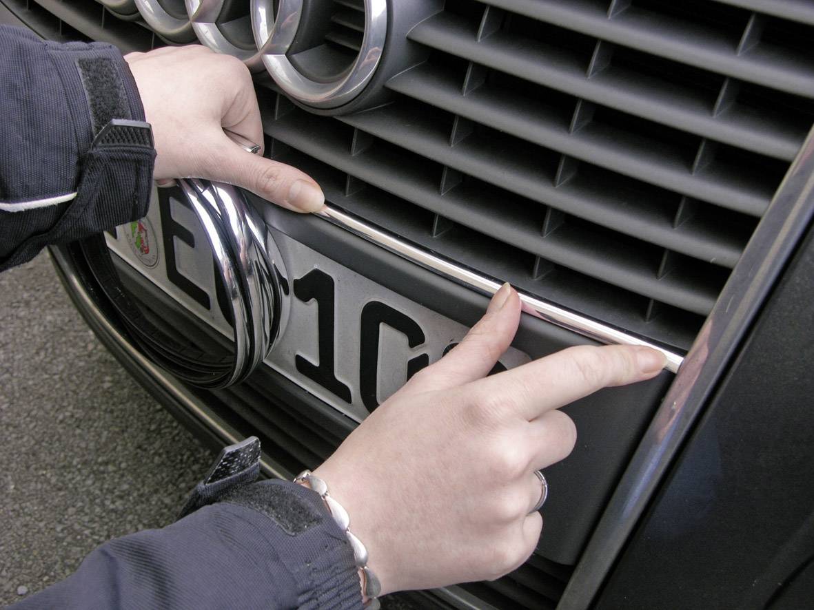 Osoba upevňuje oválný, chromovaný předmět na přední straně automobilu poblíž registrační značky.