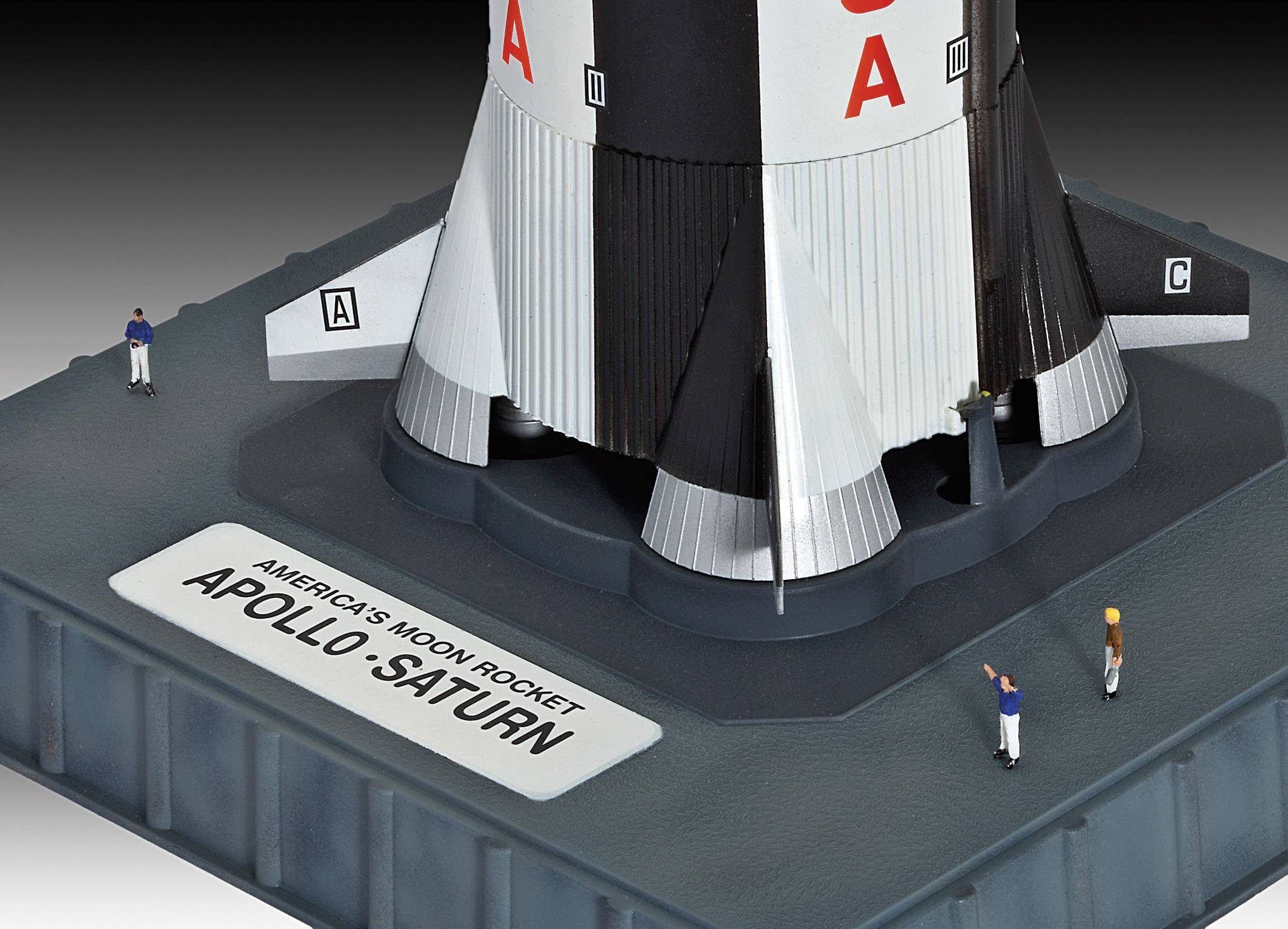 Model rakety Apollo-Saturn. Tři figury stojí kolem základny modelu, který je umístěn na platformě s popiskem.