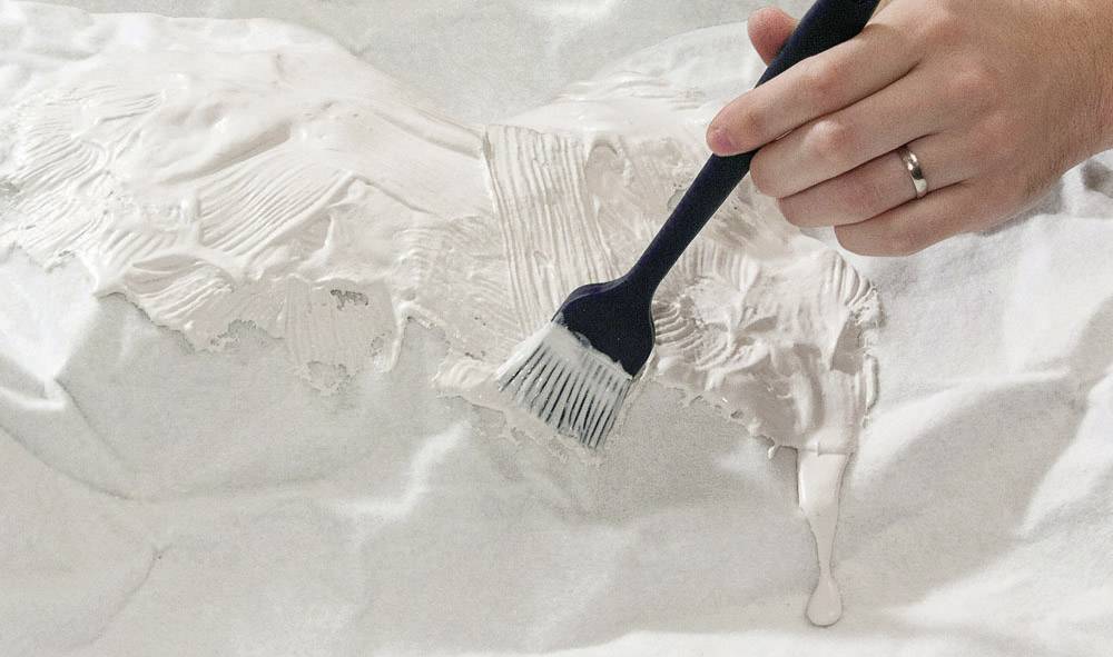 Ruka s prstenem drží štětec a nanáší bílou barvu na strukturovaný povrch.