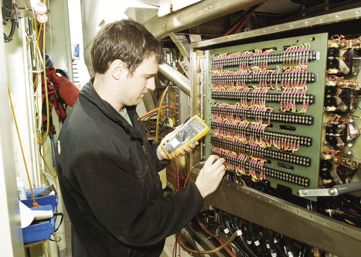 Technik kontroluje elektrické spoje multimetrem na rozvodné desce v technické místnosti.
