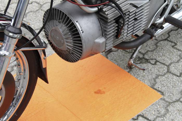 "Motorka s olejovou skvrnou" zobrazuje motocyklový motor na oranžové podložce na dlažebních kostkách. Je patrná malá olejová skvrna.