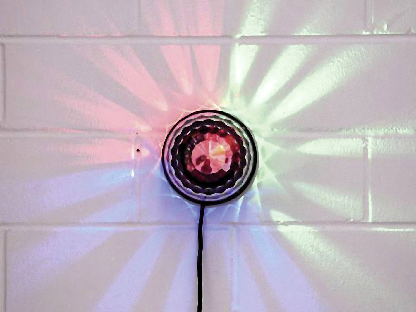 Kulaté nástěnné světlo s barevným osvětlením; promítá červené, zelené a modré paprsky na bílou cihlovou stěnu.