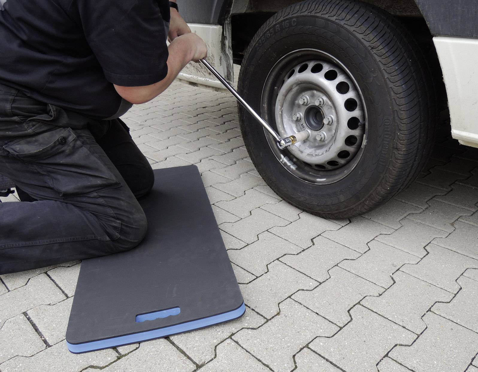 Muž klečí na dlažebních kostkách a pomocí klíče vymění kolo automobilu. Černá podložka chrání jeho kolena.