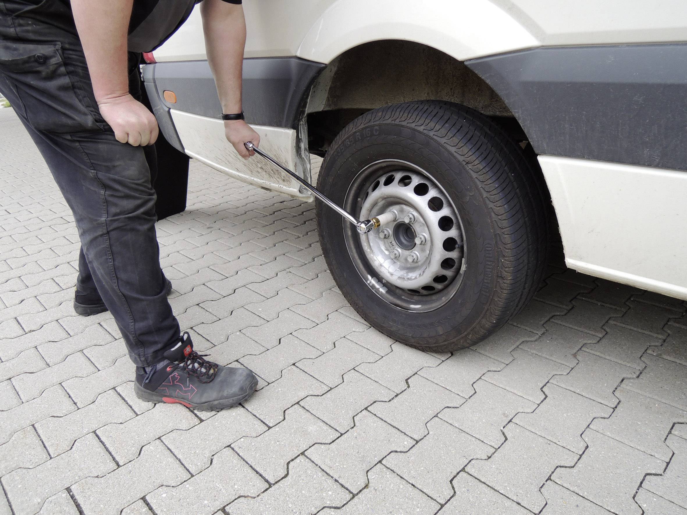 Muž vymění kolo na bílém dodávkovém voze. Používá křížový klíč k povolení šroubů, zatímco vozidlo stojí na dlážděném povrchu.