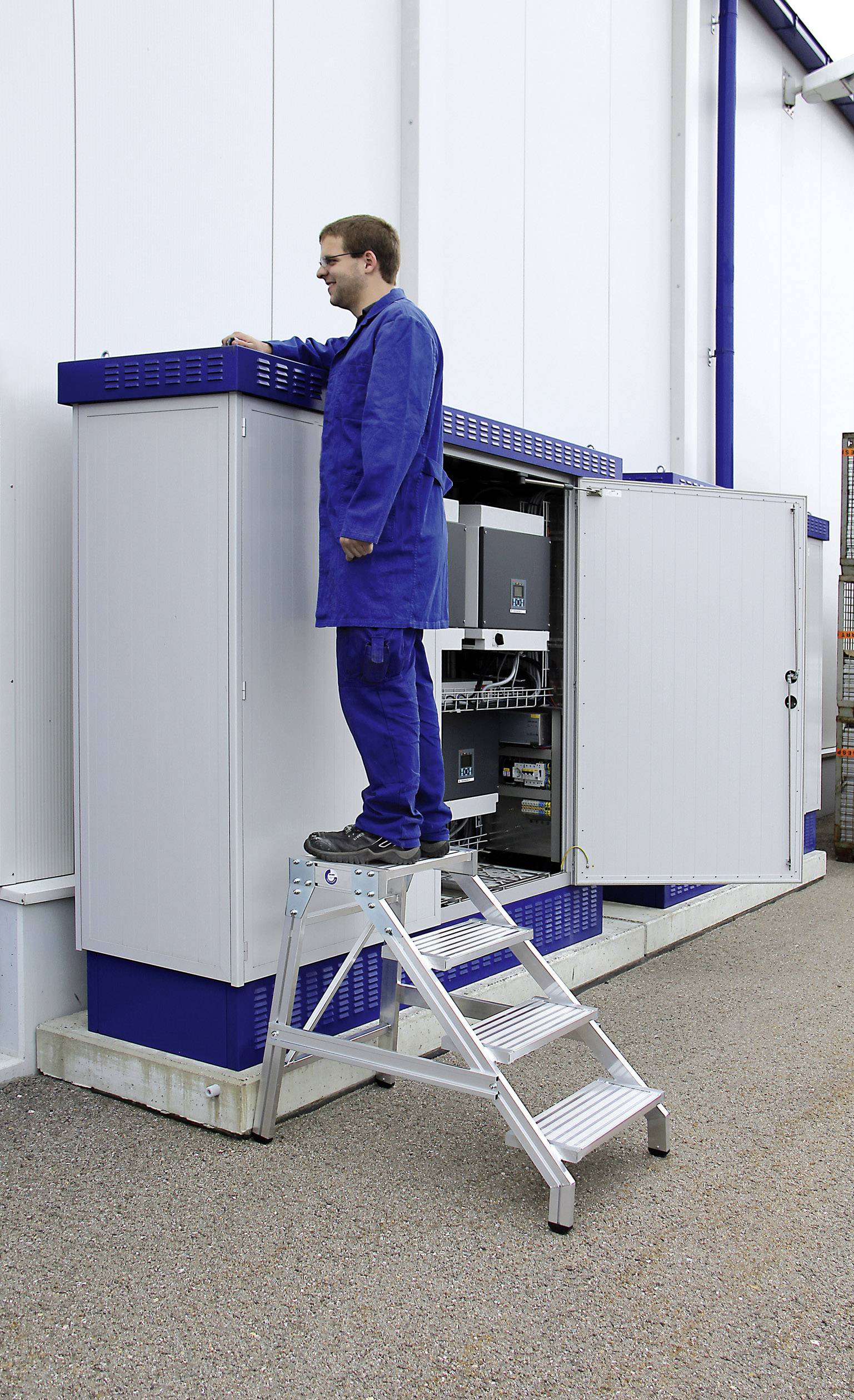 Muž stojí na žebříku a pracuje na otevřené rozvodné skříni. Má na sobě modrý pracovní oděv. Rozvodná skříň je připevněna na stěně.