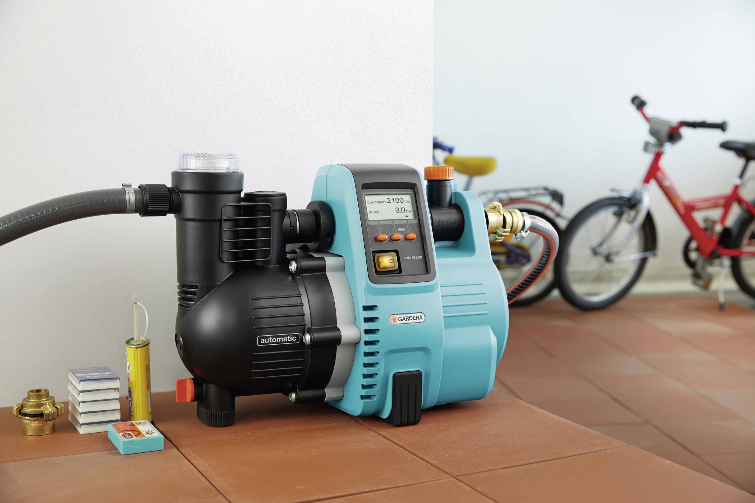 Zahradní domácí vodárna Gardena na terase, s kolem v pozadí. Zobrazuje vodní čerpadlo s LCD displejem, potrubím a příslušenstvím.