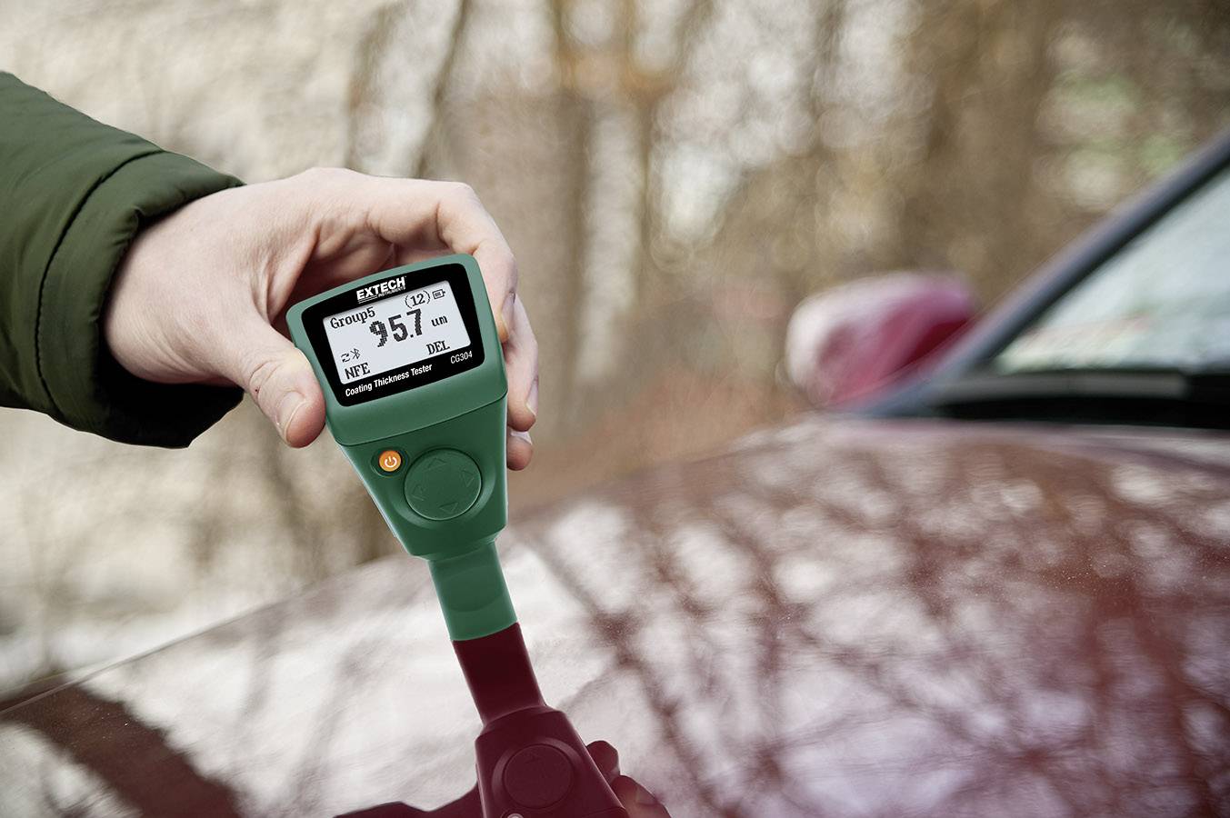 Jedna ruka drží digitální měřicí přístroj, který ukazuje tloušťku lakové vrstvy automobilu na 95,7 mikrometrů.