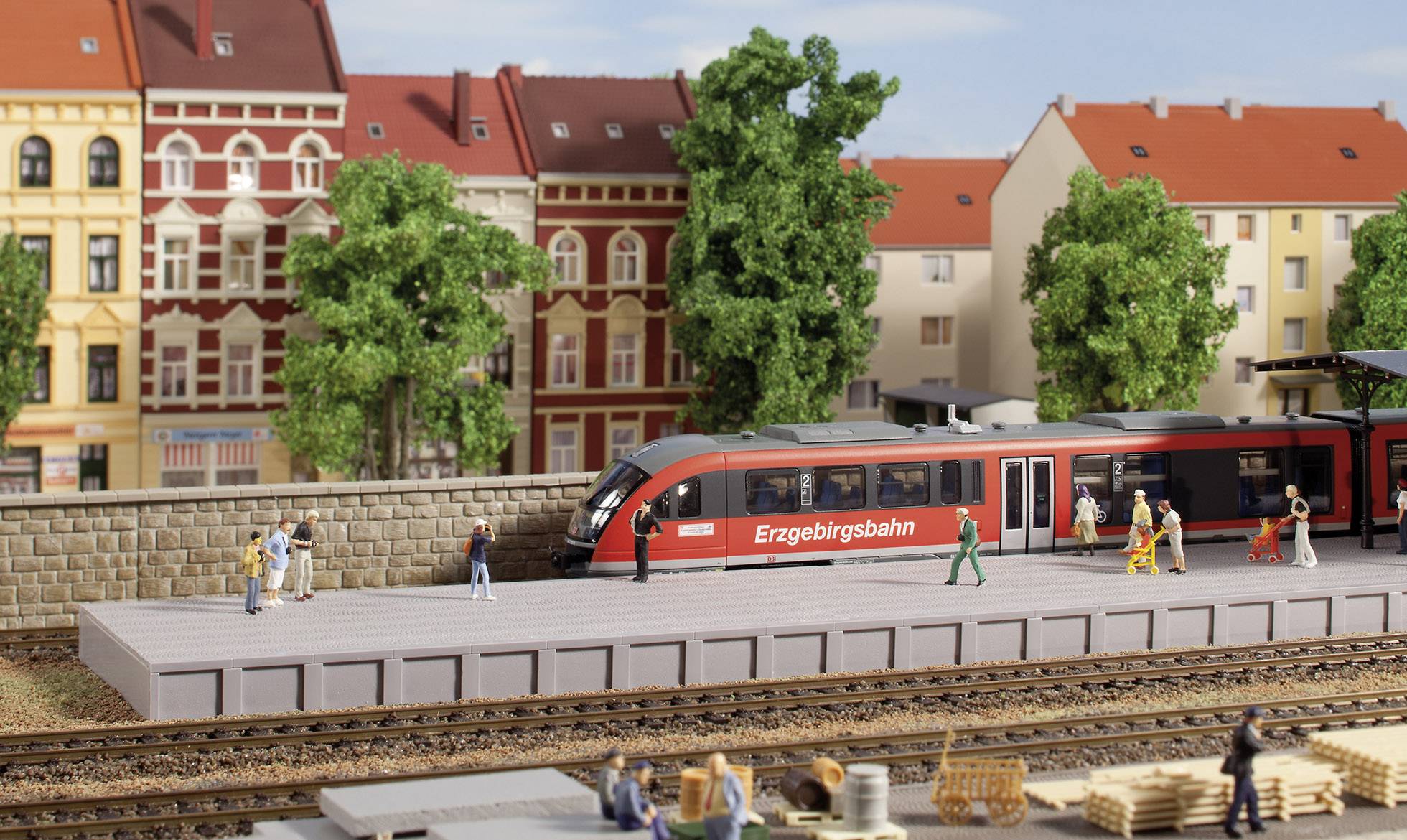 Model železnice zobrazuje červený vlak na nástupišti ve městě s historickými budovami a stromy.