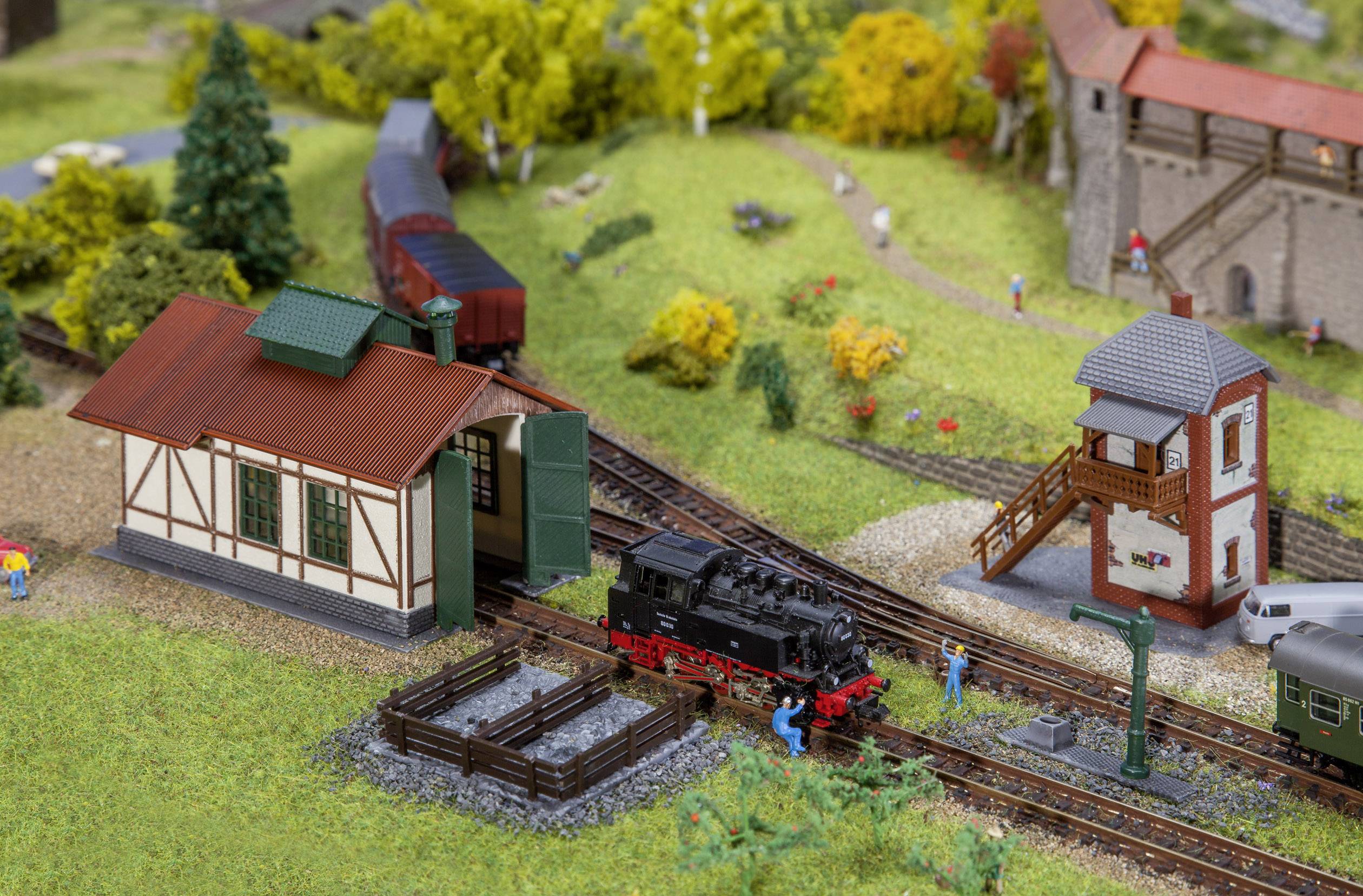 Detailní modelová železnice s malými budovami, vlaky a figurkami. Parní lokomotiva jede vedle nádražní budovy přes venkovskou krajinu.