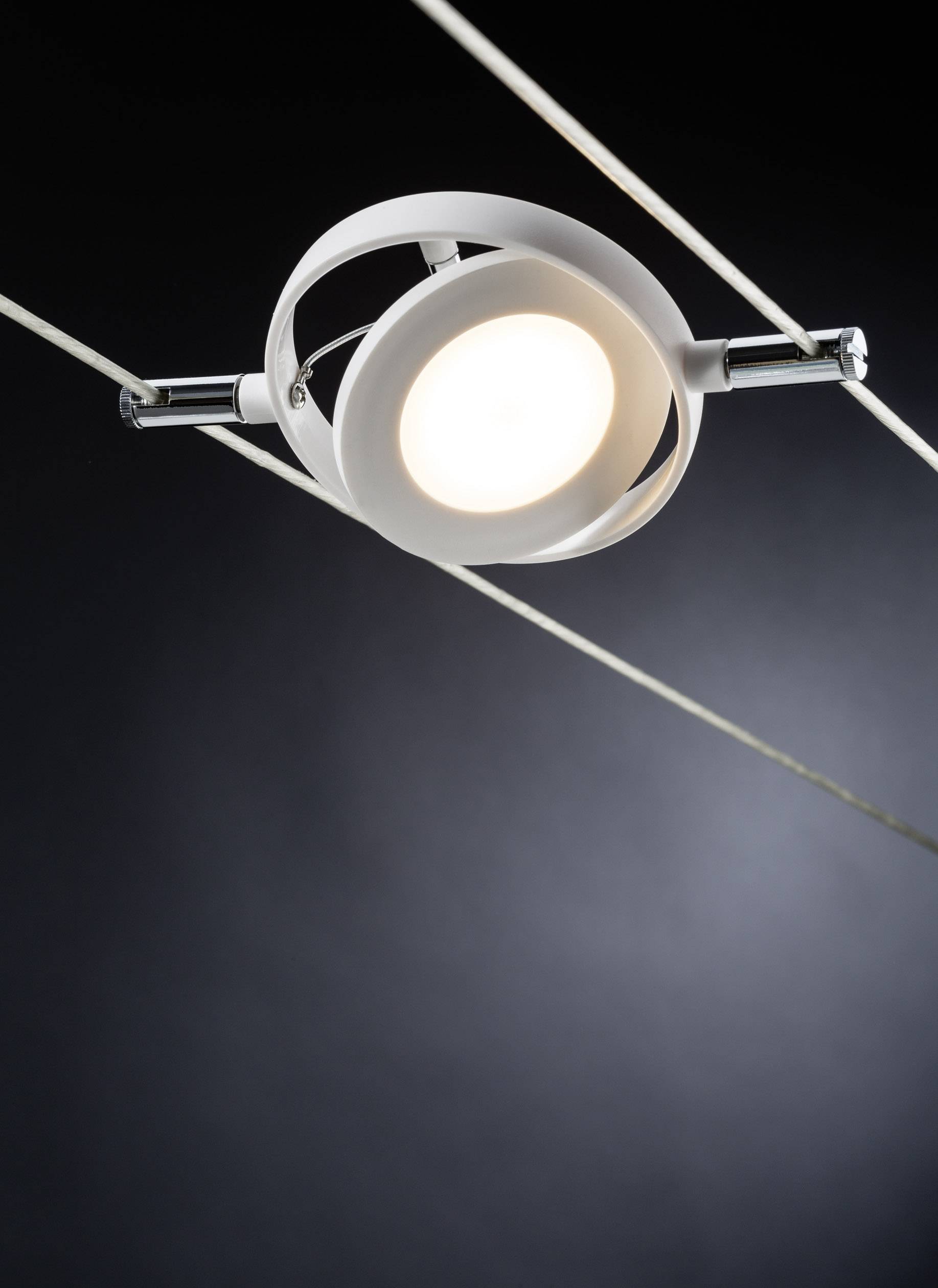 Moderní lampa zavěšená na lanech září v temné místnosti. Je kulatá, kovová, s jasným světlem uprostřed.