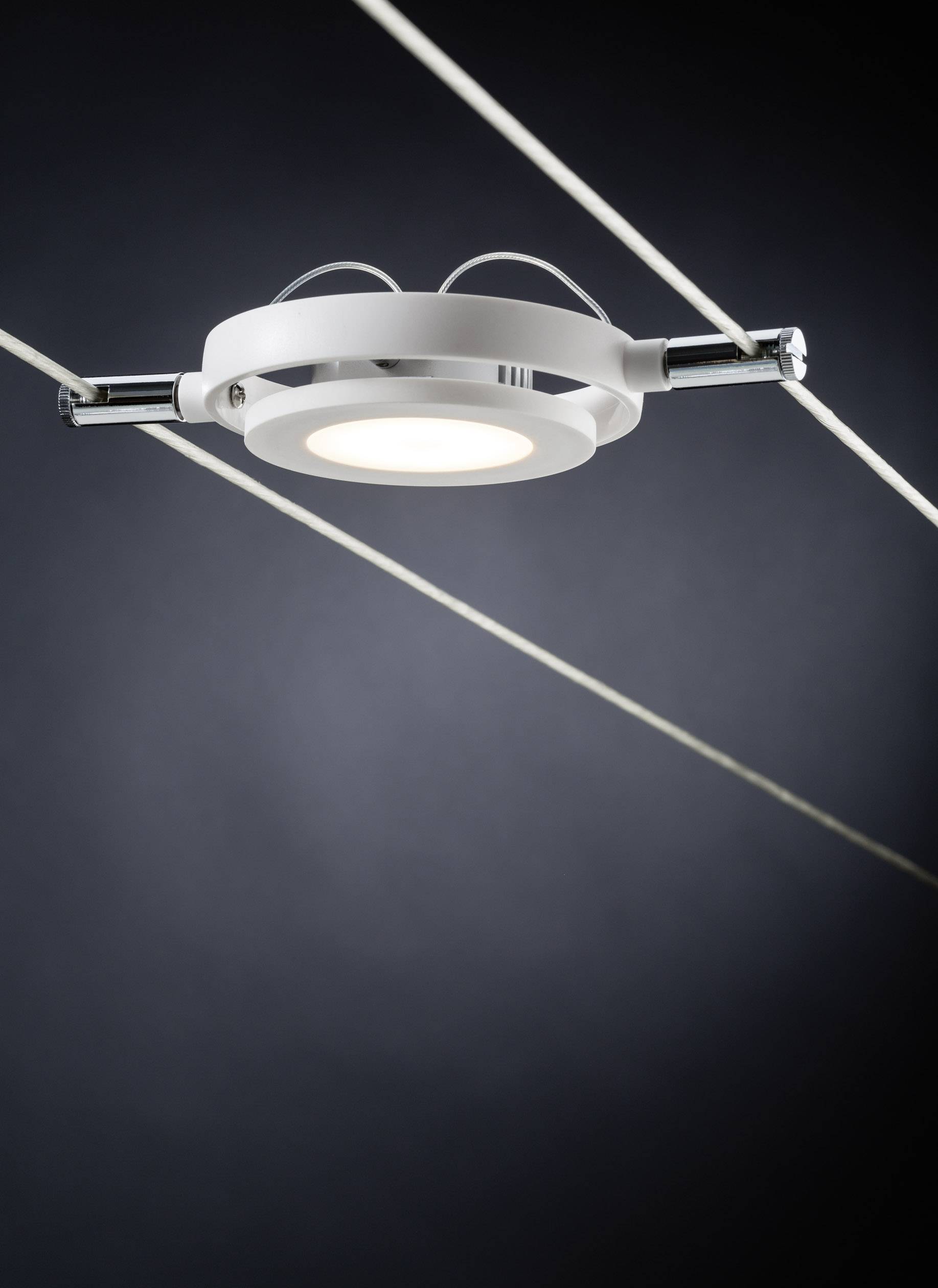 Moderní kulatá stropní lampa z kovu visí na dvou rovnoběžně napnutých kabelech před tmavým pozadím.