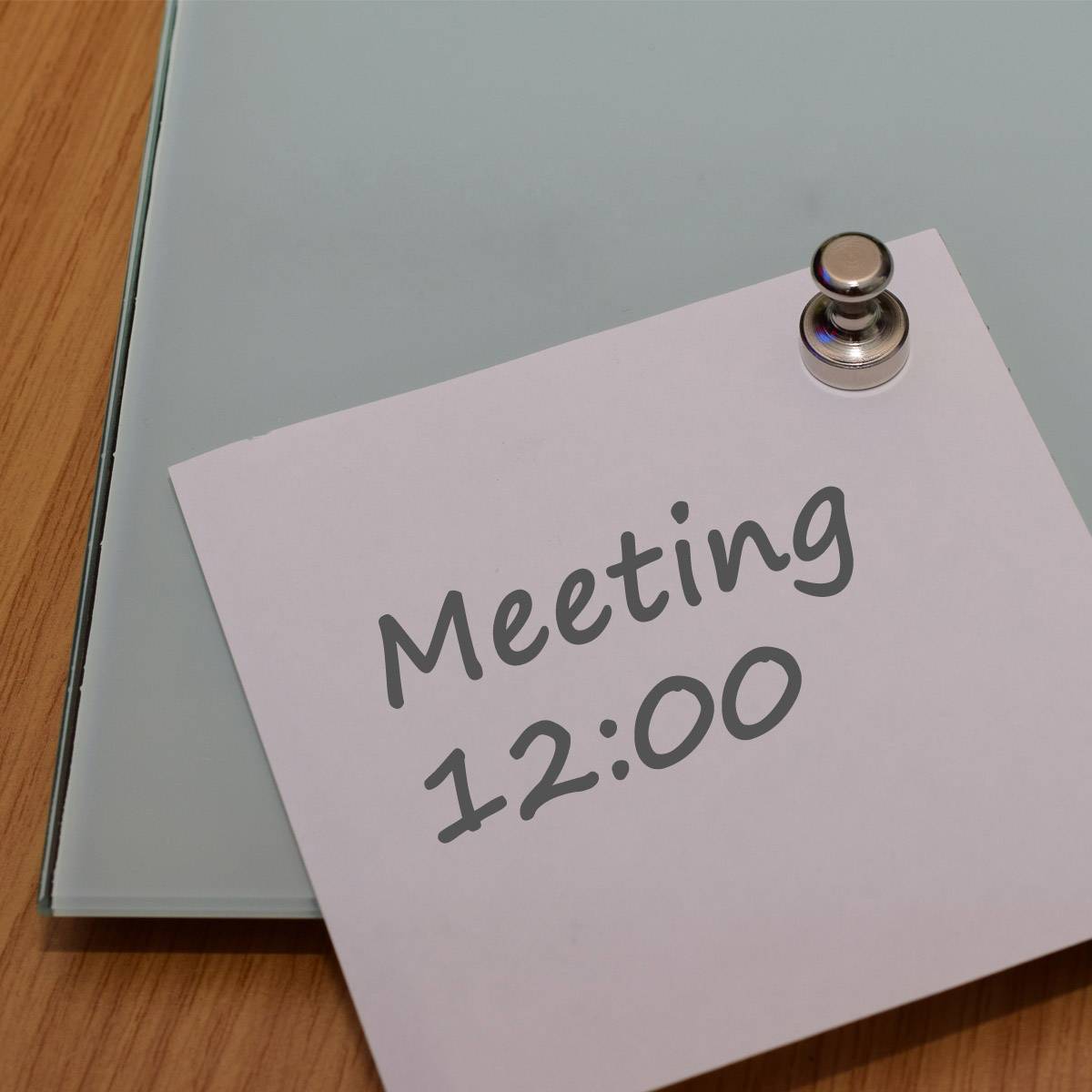Bílá poznámka na psacím stole, připevněná svorkou, informuje o schůzce ve 12:00 hodin.