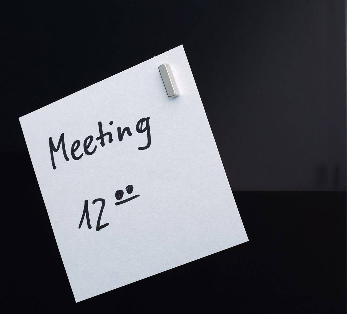 Bílý poznámkový lístek upevněný magnetem zobrazuje rukou psanou poznámku 'Schůzka 12:00' na tmavém povrchu.