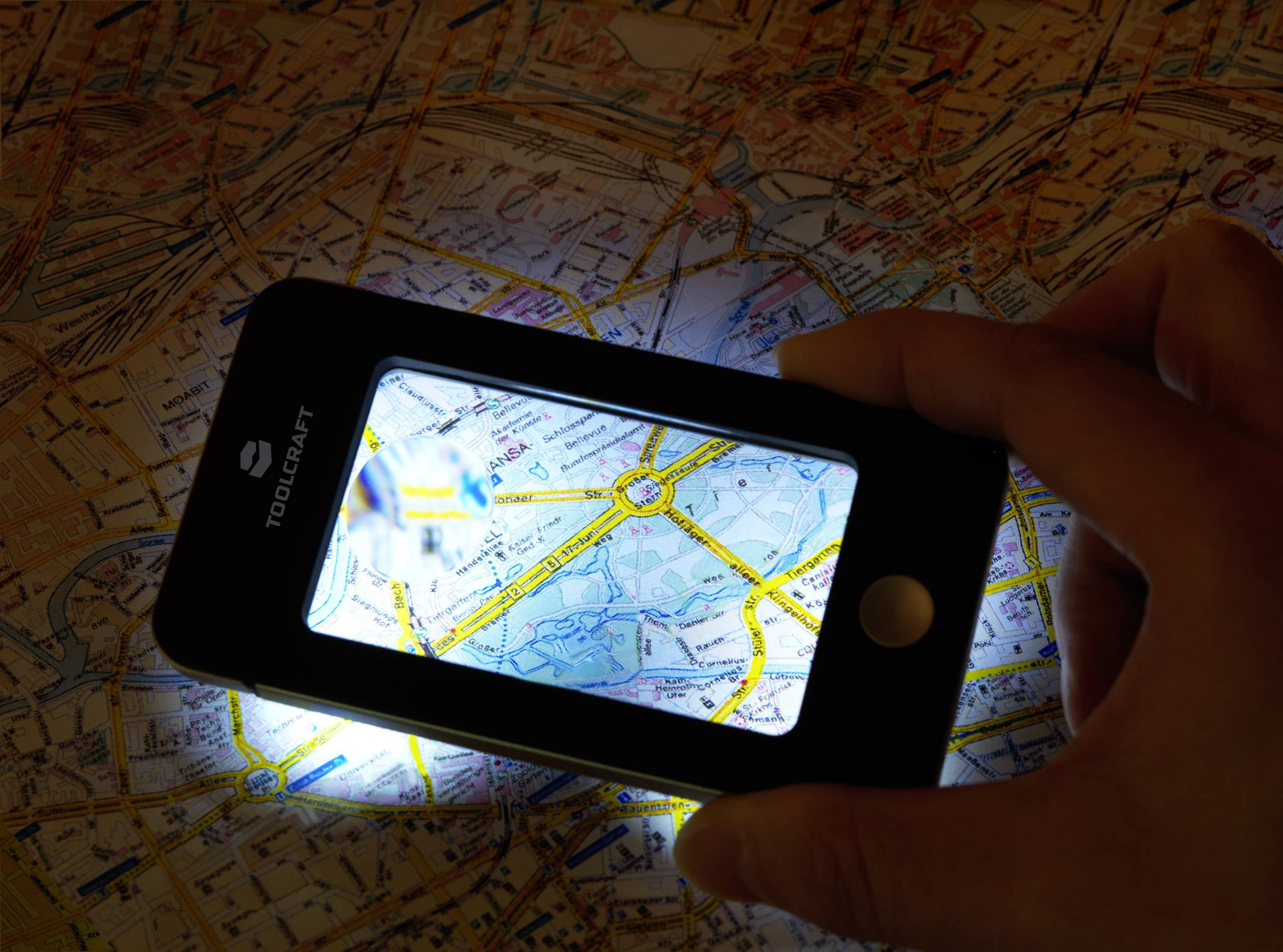 Jedna ruka drží osvětlenou lupu nad mapou města, aby zvětšila detaily. Mapa zřetelně ukazuje ulice a místní názvy.