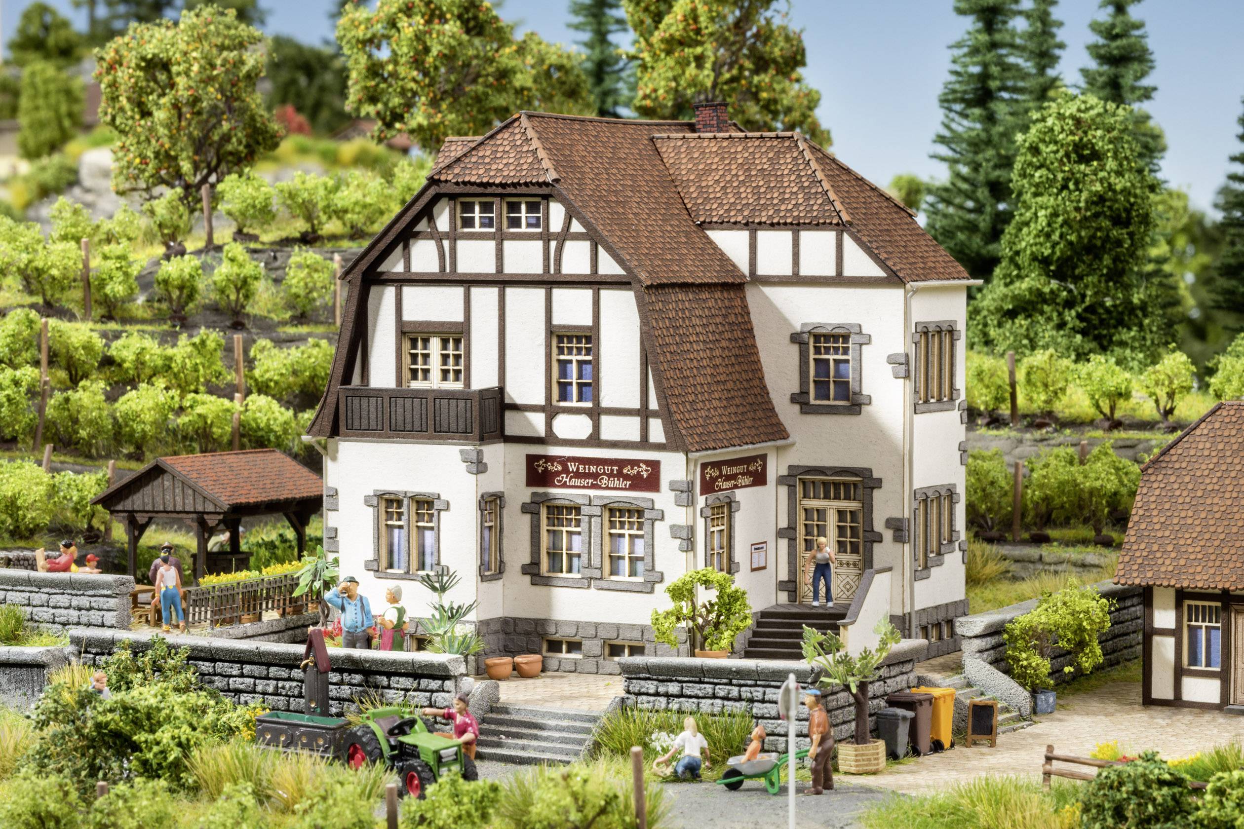Miniaturní model hrázděného domu s vinicemi v pozadí. Lidé jsou rozmístěni kolem domu, někteří jsou ponořeni do hovoru.