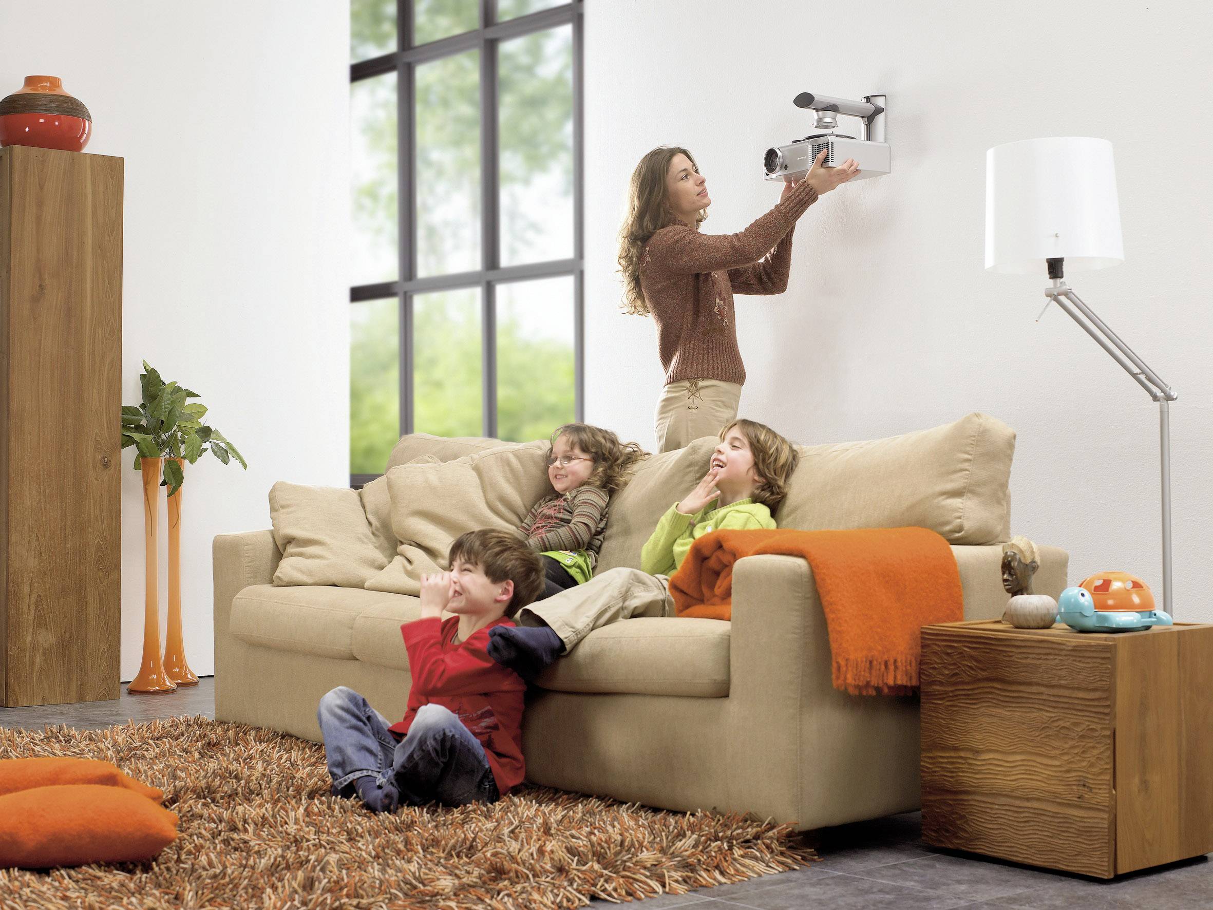 Žena instaluje projektor na stěnu, zatímco tři děti sedí na béžové pohovce a přihlížejí.