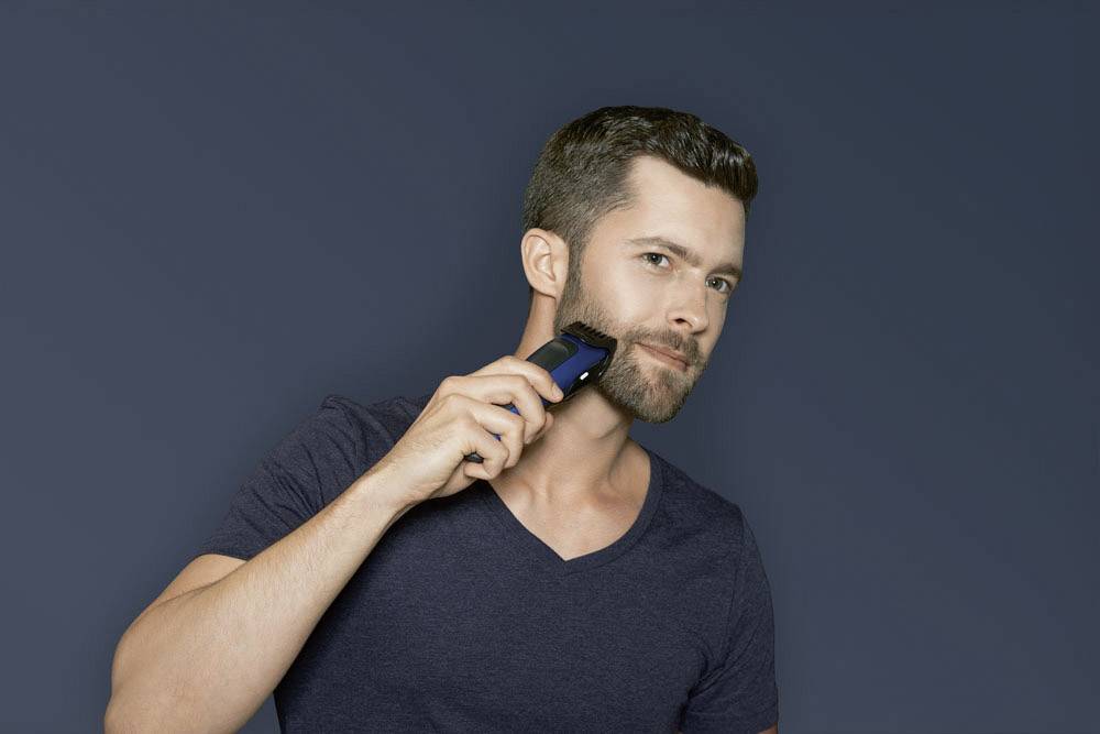 Muž se holí elektrickým holicím strojkem před tmavě modrým pozadím.