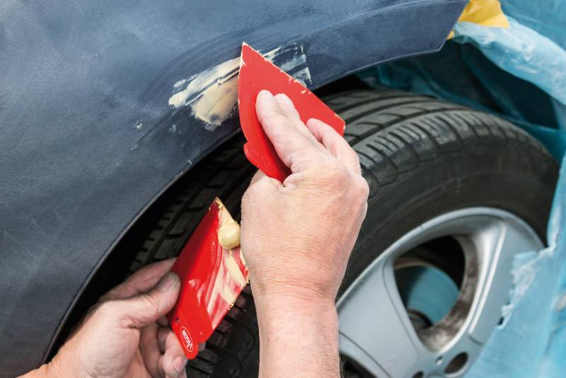 Jedna osoba opravuje karoserii automobilu, nanáší stěrkovou hmotu na blatník červenými stěrkami, aby opravila poškození.