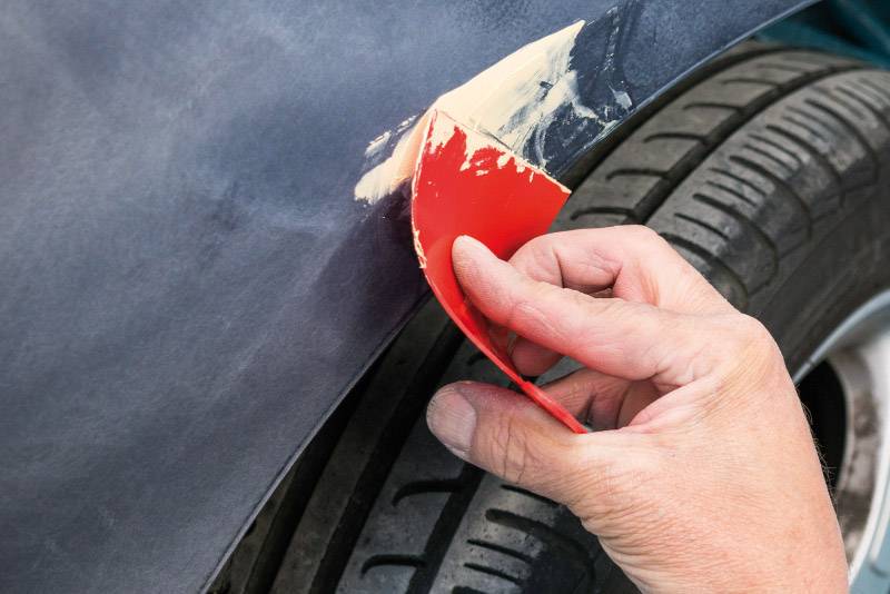 Ruka roztírá tmelicí hmotu na škrábanec karoserie pomocí červeného nářadí. Detailní záběr ukazuje precizní práci na karoserii automobilu.