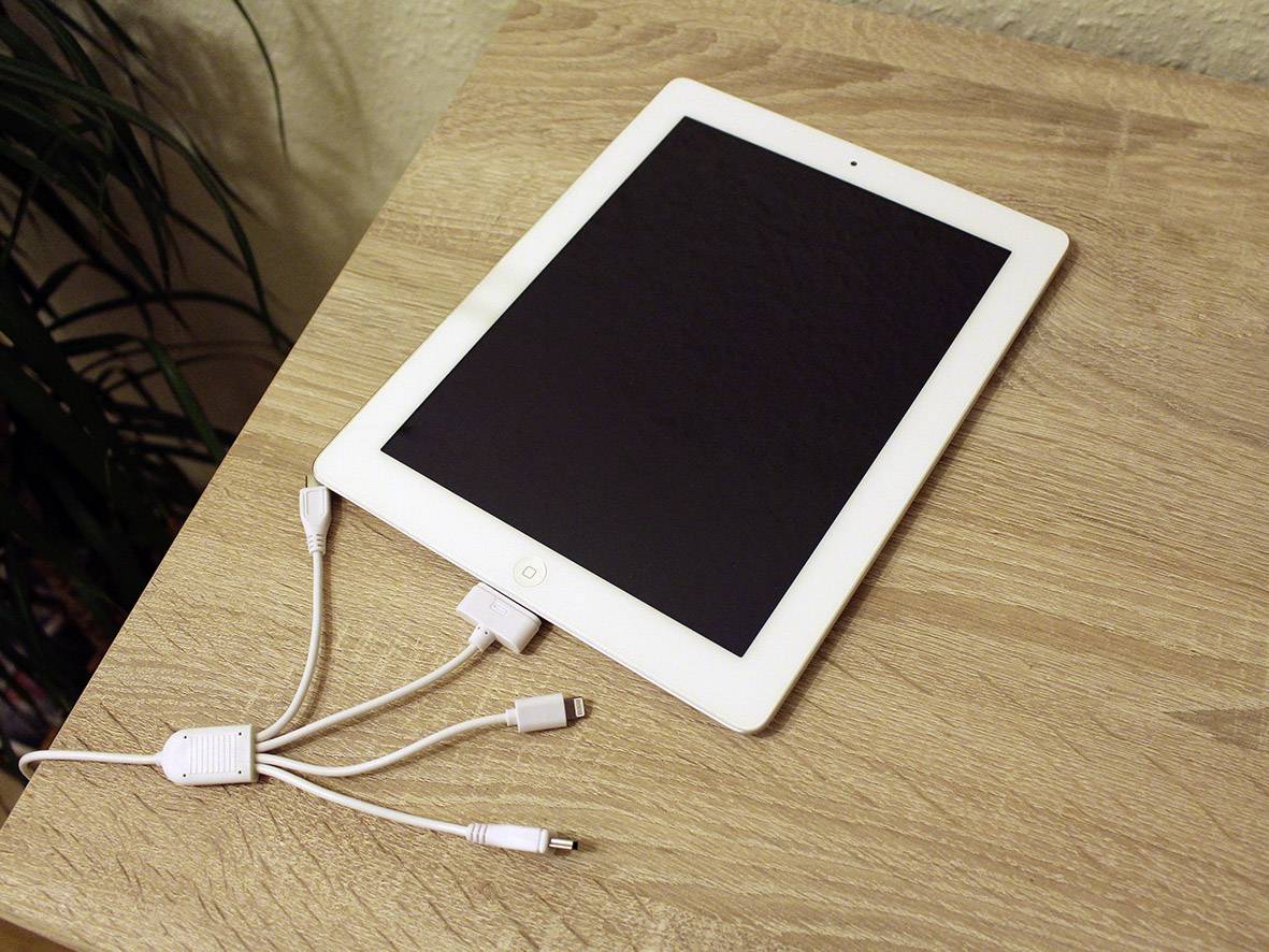 Bílý tablet s černou obrazovkou na dřevěném stole, připojený několika bílými kabely. V pozadí je vidět rostlina.