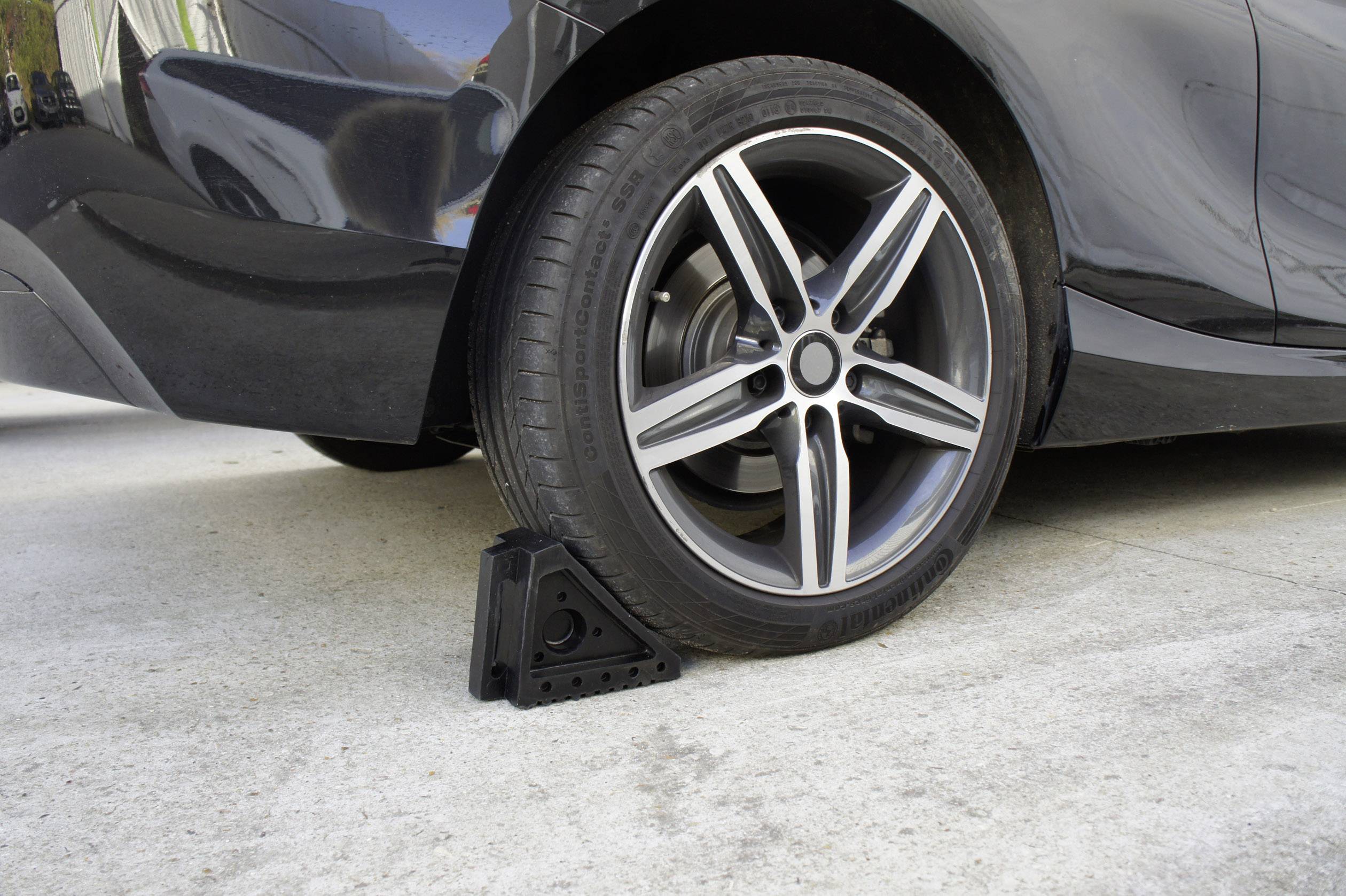 Černé auto s brzdným klínem před pneumatikou, který zajišťuje vozidlo na betonovém povrchu.