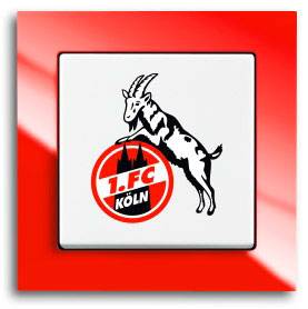 Kozí beran stojí na míči s logem '1. FC Köln'. Symbolizuje fotbalový klub z Kolína nad Rýnem.