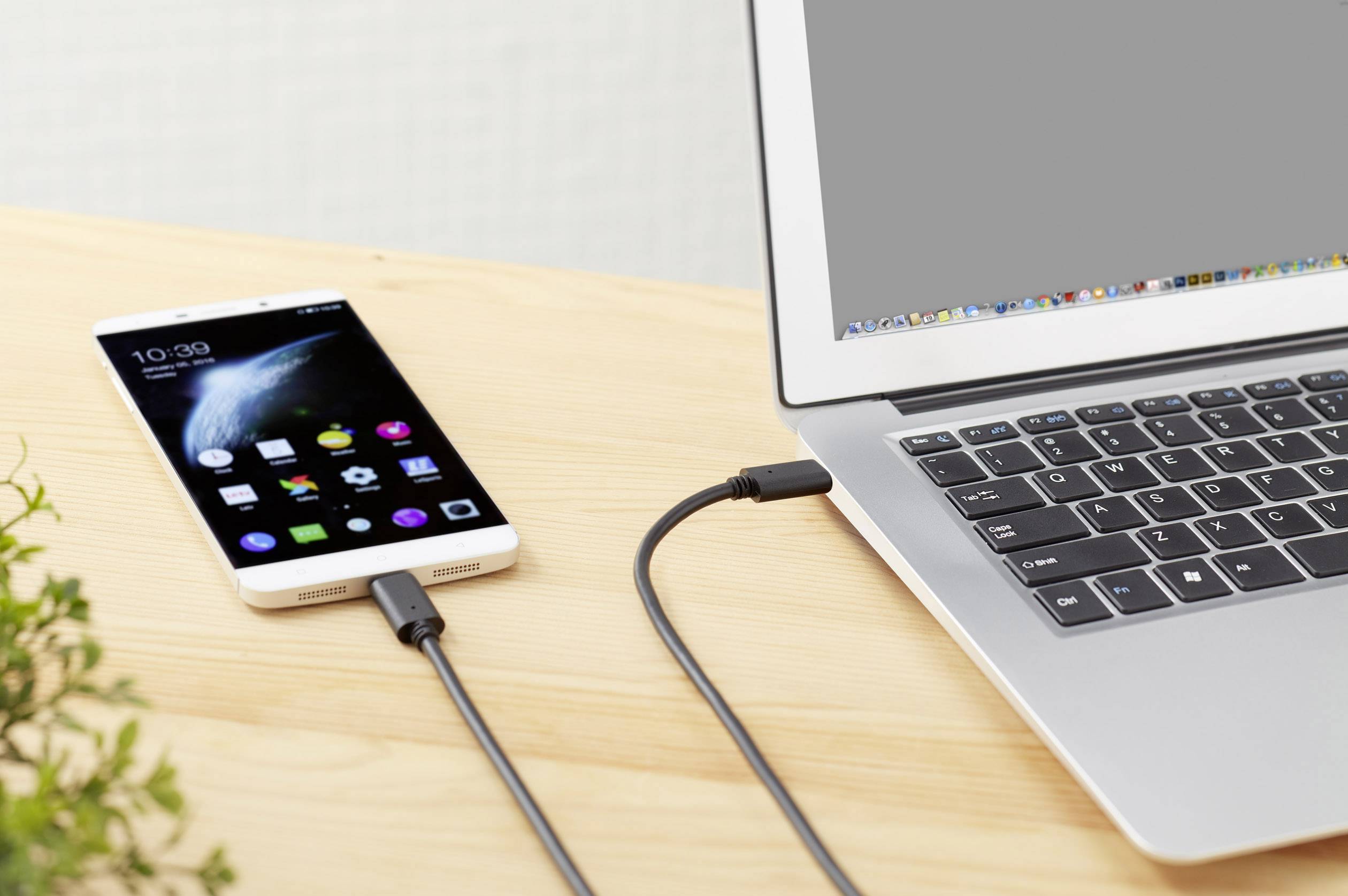 Smartphone připojený k laptopu přes USB kabel, umístěný na dřevěném stole. Obrazovka laptopu je vypnutá.