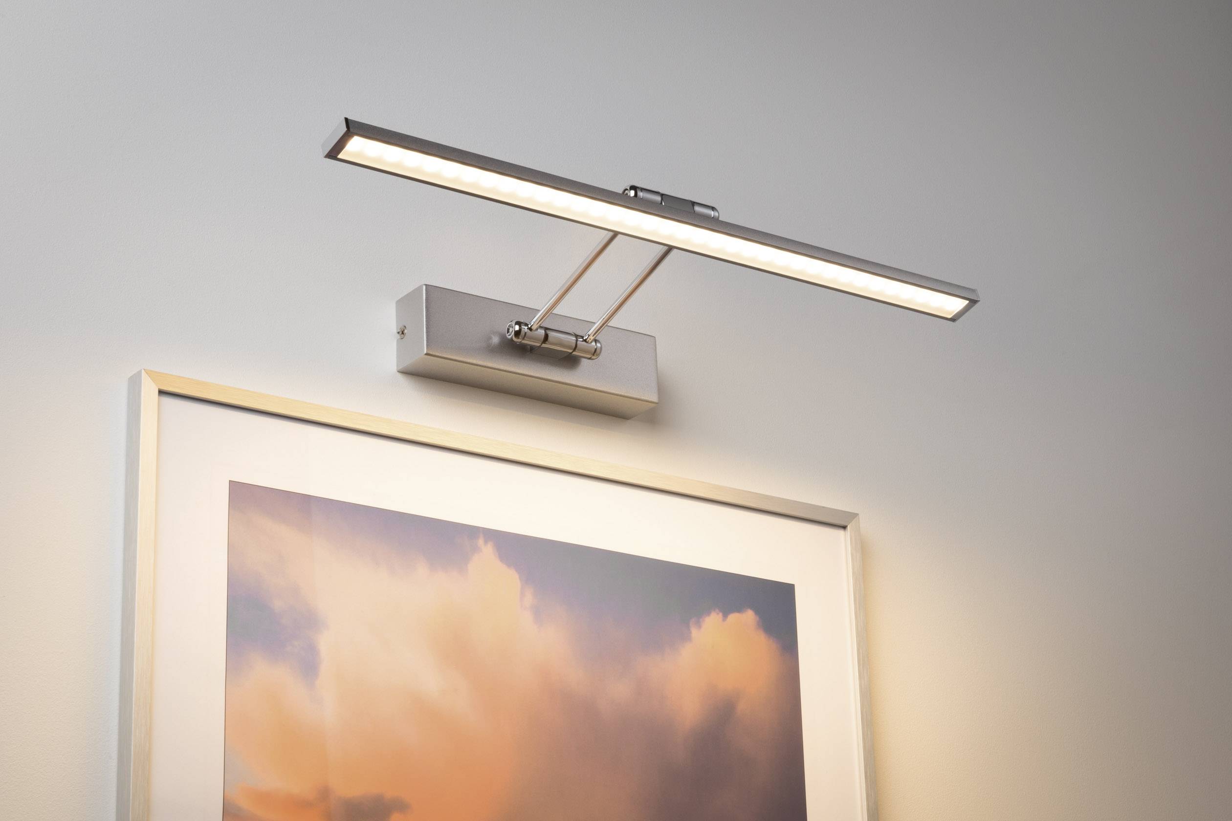 Štíhlá nástěnná lampa osvětluje obraz s oranžovými mraky, který visí na šedé stěně.