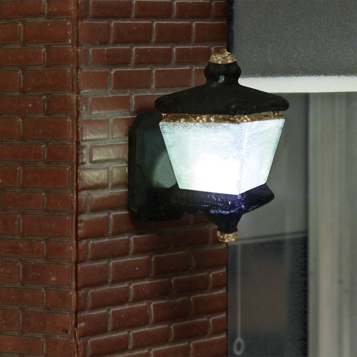 Nástěnná lampa s matným sklem a kovovým rámečkem zavěšená na cihlové zdi, která v soumraku vyzařuje jemné světlo.