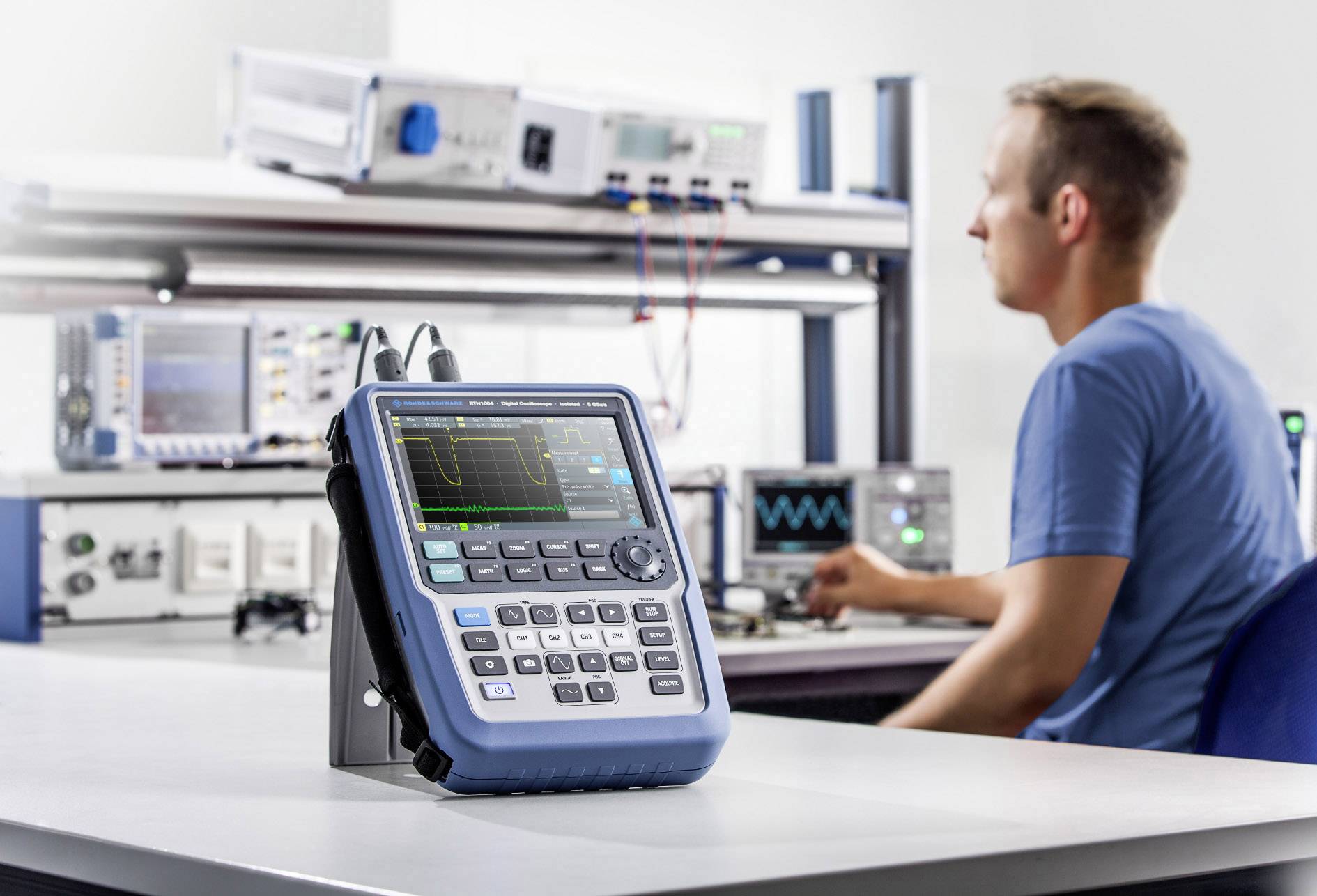 Technik sedí v laboratoři a pracuje s osciloskopem umístěným na stole. V pozadí jsou vidět další elektronické přístroje.