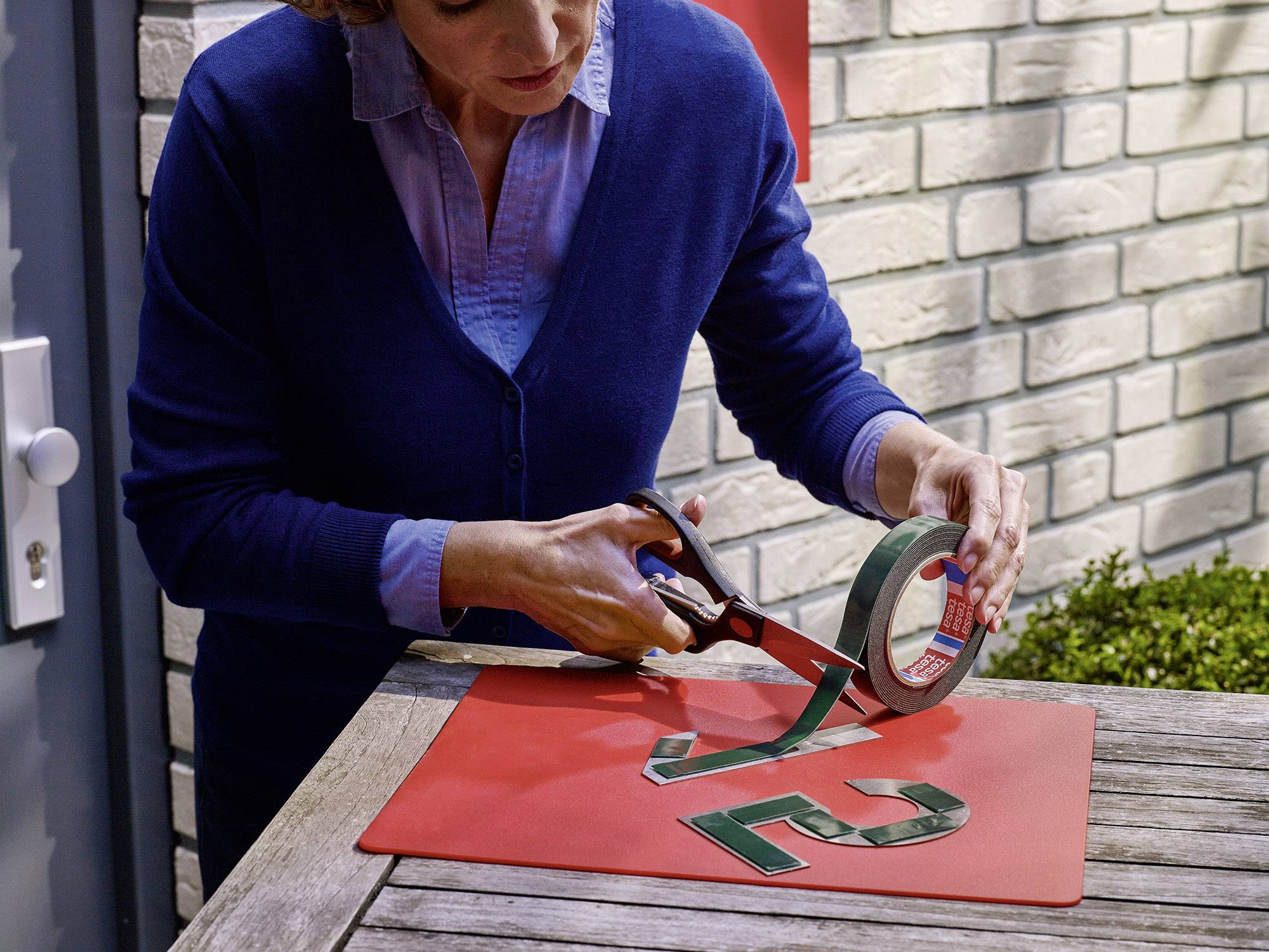 Jedna osoba stříhá lepicí pásku nůžkami na dřevěném stole. Na stole leží zelené číslovky, červená deska a v pozadí cihlová zeď.