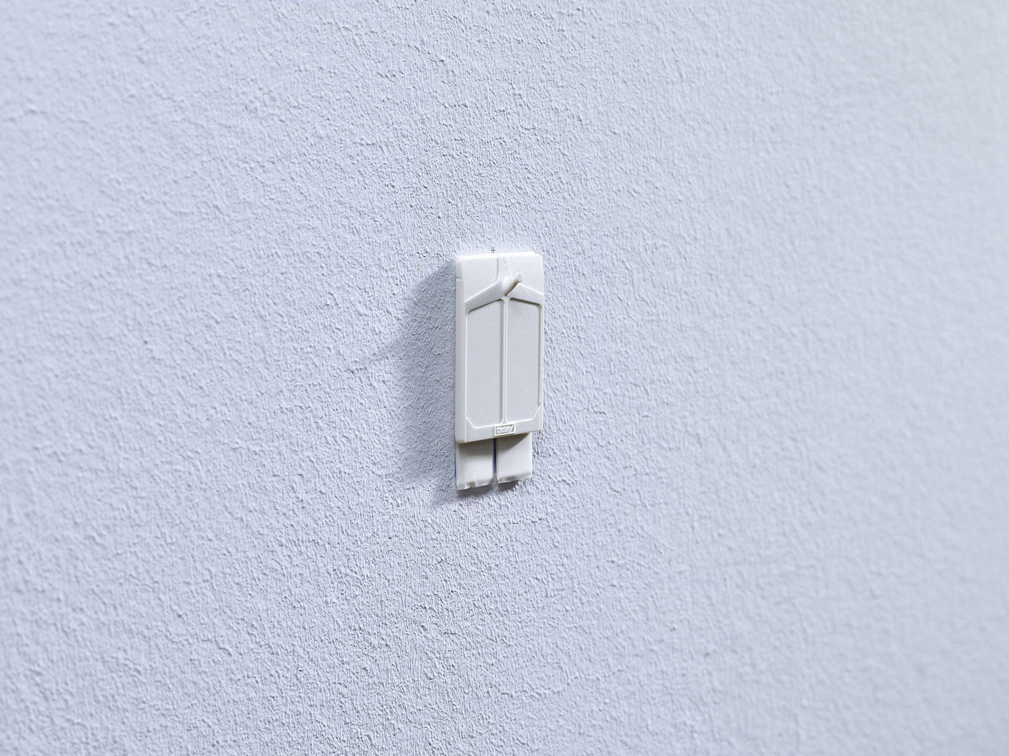 Bílá plastová lišta se háčky na šedé zdi, připravená k zavěšení předmětu. Jednoduché, funkční designové řešení.