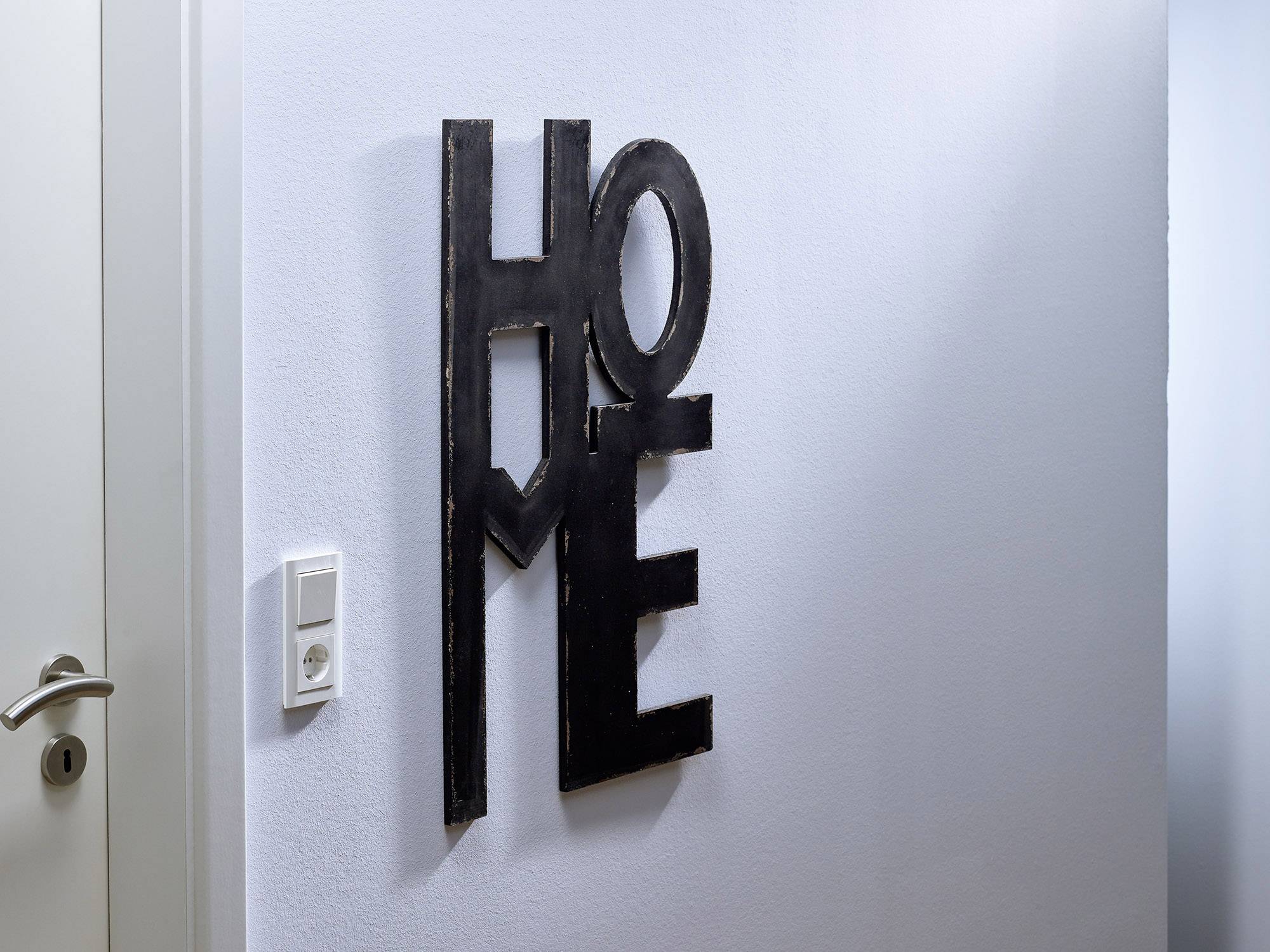 Černá písmena na bílé zdi vytvářející slovo 'HOME'. Vlevo je částečně viditelný vypínač a dveře.