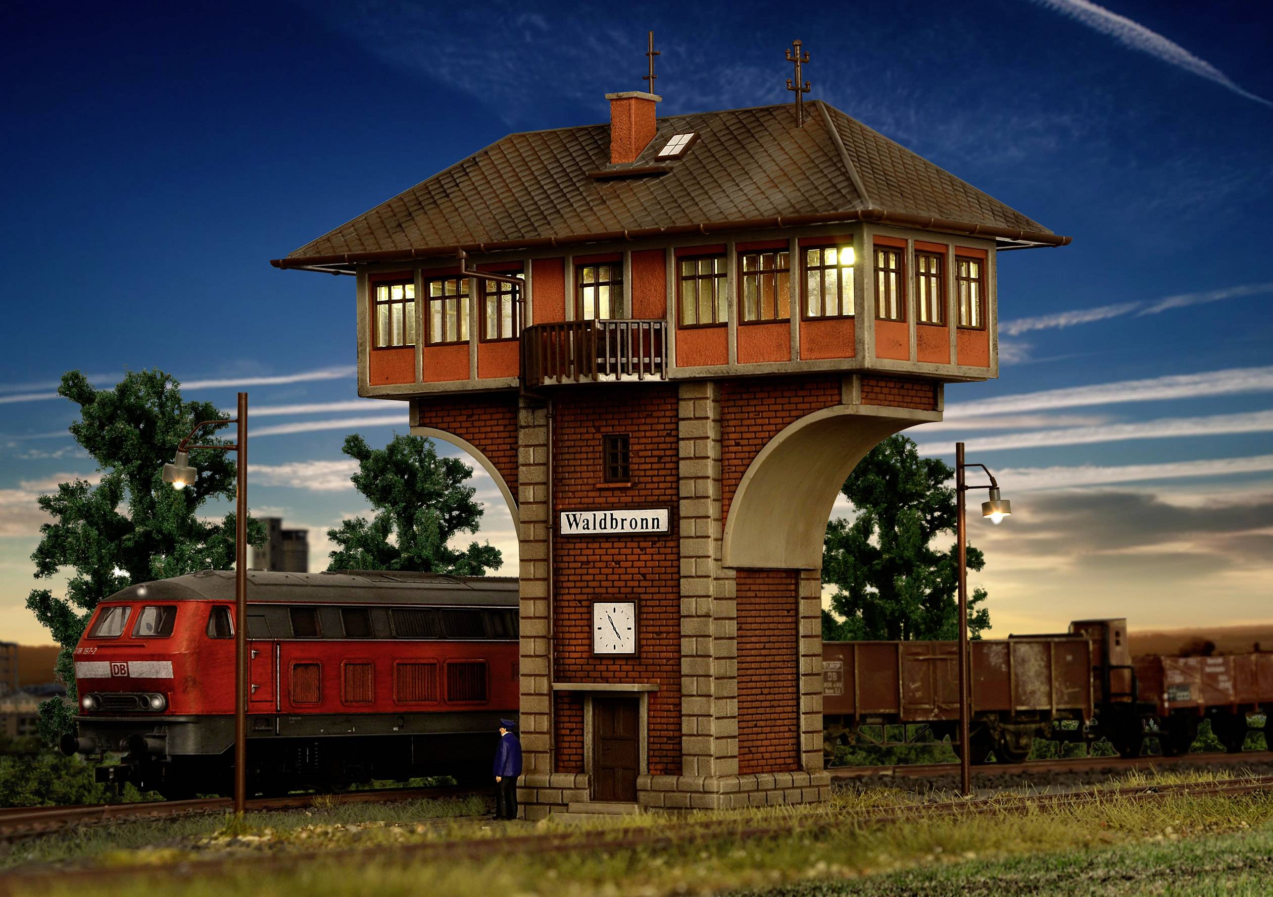 Červený model vlaku projíždí kolem strážního domku 'Waldbronn'. V pozadí jsou stromy a osvětlená večerní obloha.