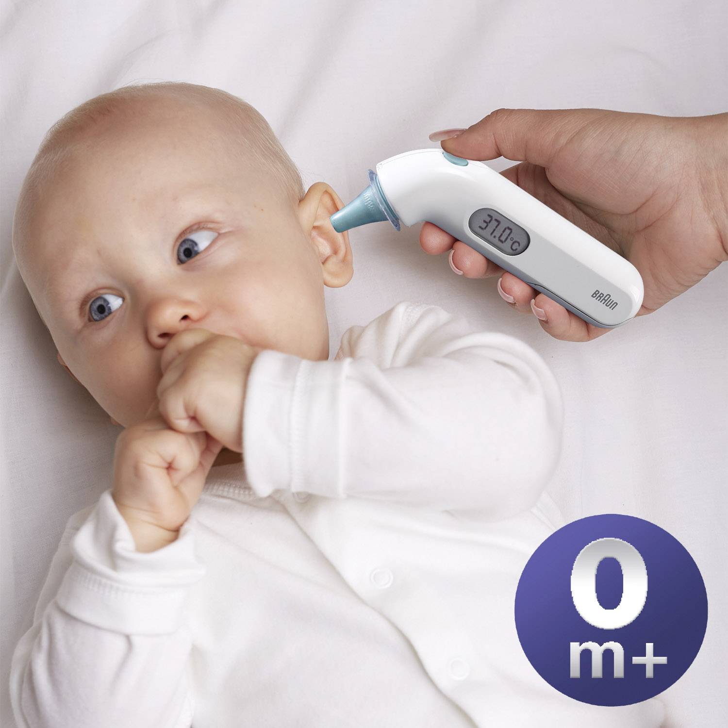Miminko leží na posteli, zatímco ruka používá digitální ušní teploměr, který ukazuje 37,0°C.