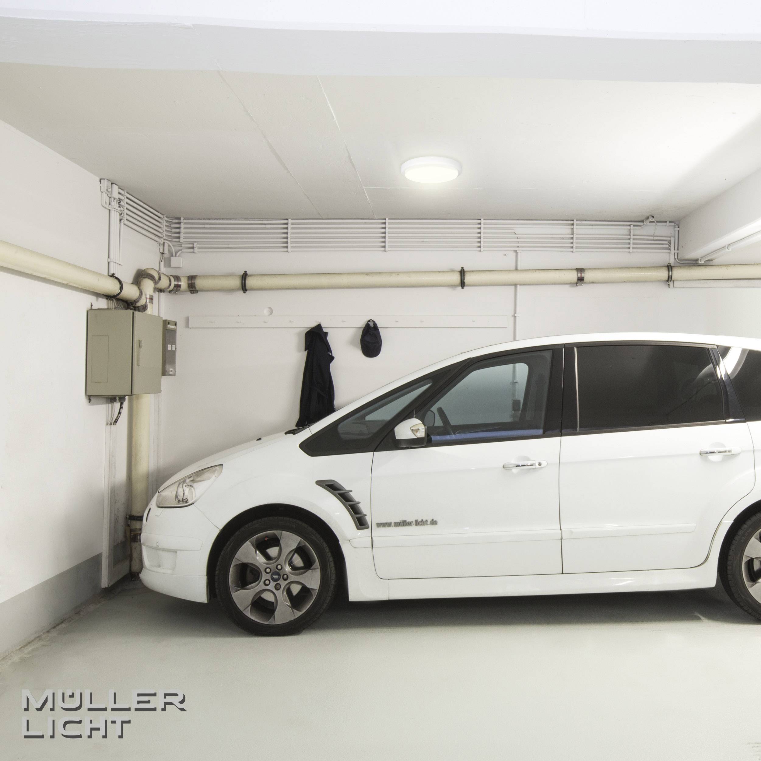 Bílé auto je zaparkované v parkovacím domě pod slabě osvětleným stropním světlem, u elektrorozvodné skříně na stěně.