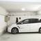 Bílé auto je zaparkované v parkovacím domě pod slabě osvětleným stropním světlem, u elektrorozvodné skříně na stěně.