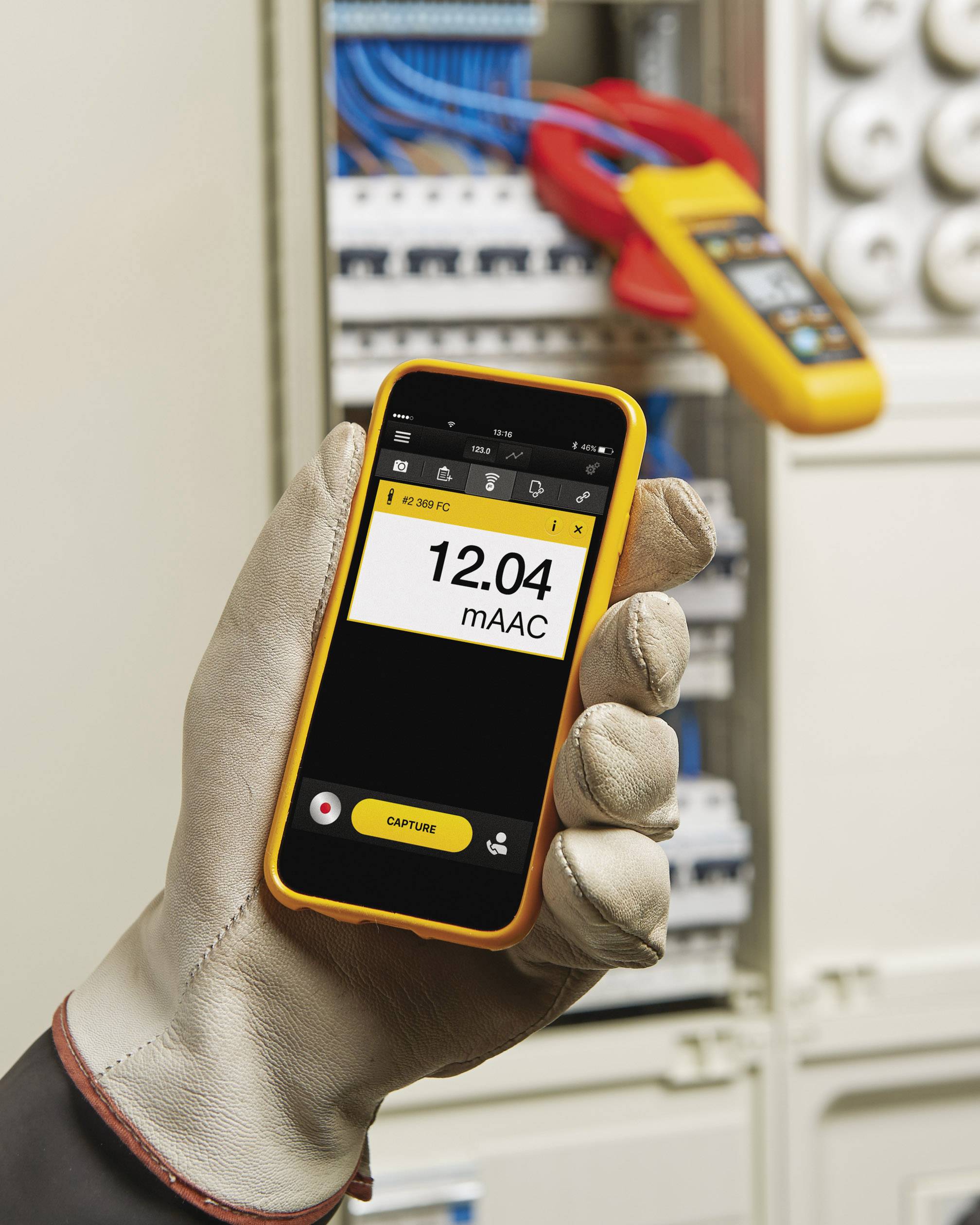 Rukavicí oblečená ruka drží smartphone s měřicí aplikací, která zobrazuje elektrickou hodnotu '12,04 mAAC'. V pozadí je otevřená rozvodná skříň.
