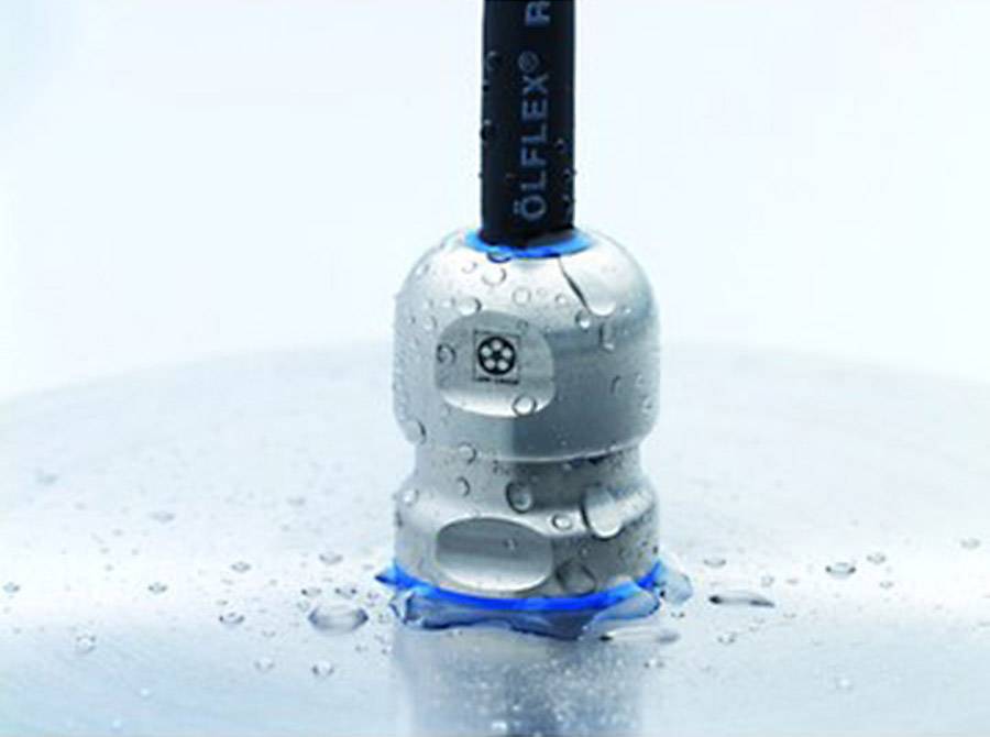Kulatý, stříbrně zbarvený kabelový průchodový prvek obklopený kapkami vody na hladkém šedém povrchu.