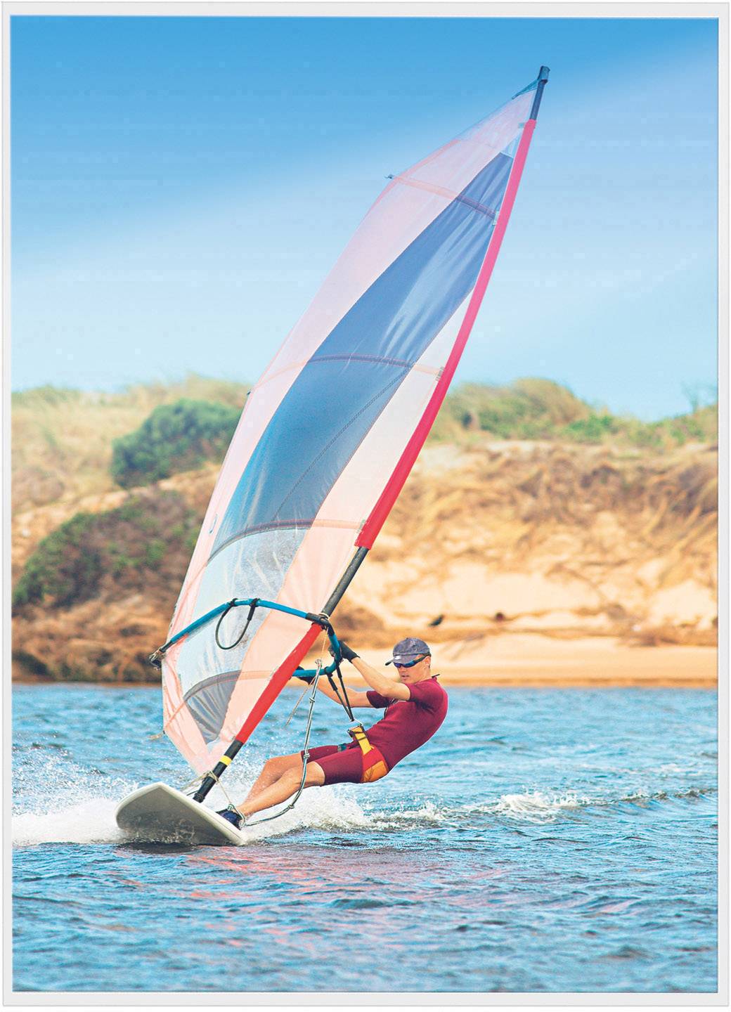 Jedna osoba surfuje na windsurfovacím prkně přes vodu s barevnou plachtou a malebným, zalesněným pobřežím v pozadí.