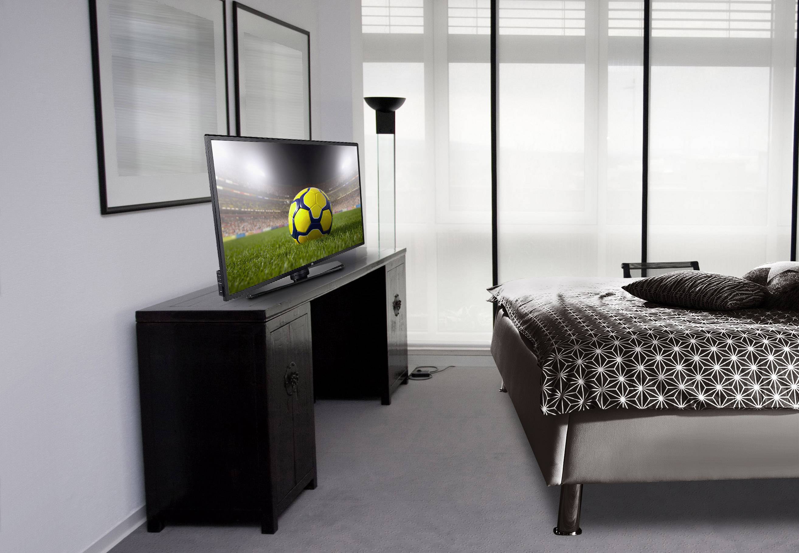 Plochý TV v moderní ložnici ukazuje fotbalový zápas. Světlá okna v pozadí, vlevo obrázky na stěně.