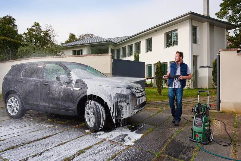 Muž myje šedé auto vysokotlakým čističem před moderním bílým domem.